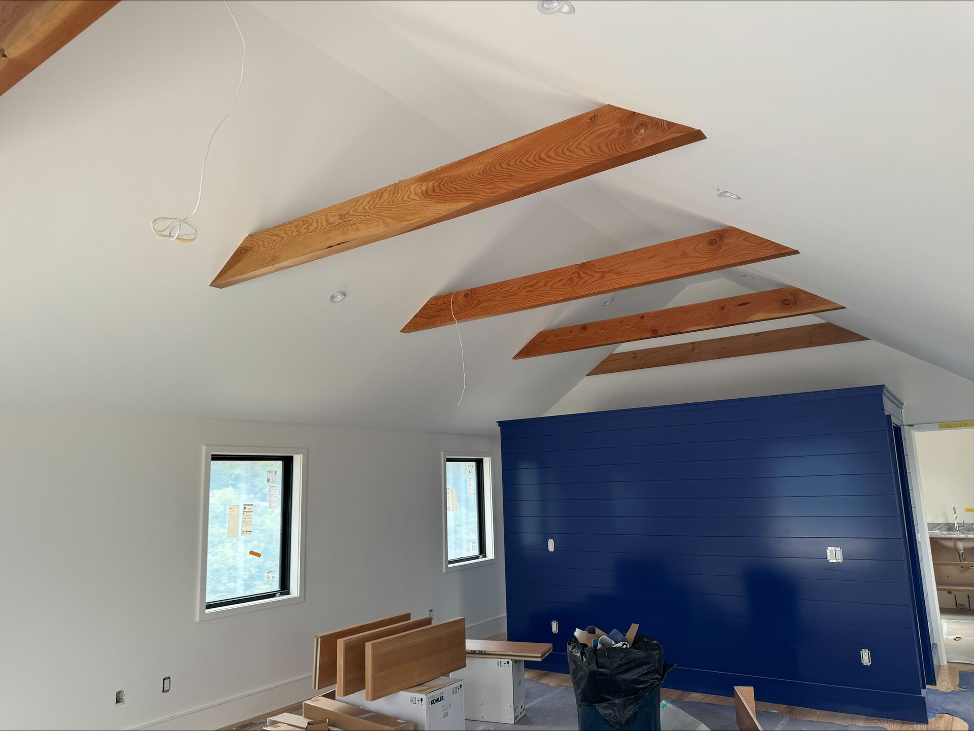 New room with sloped white ceiling, exposed wood beams, windows, and a blue accent wall under construction