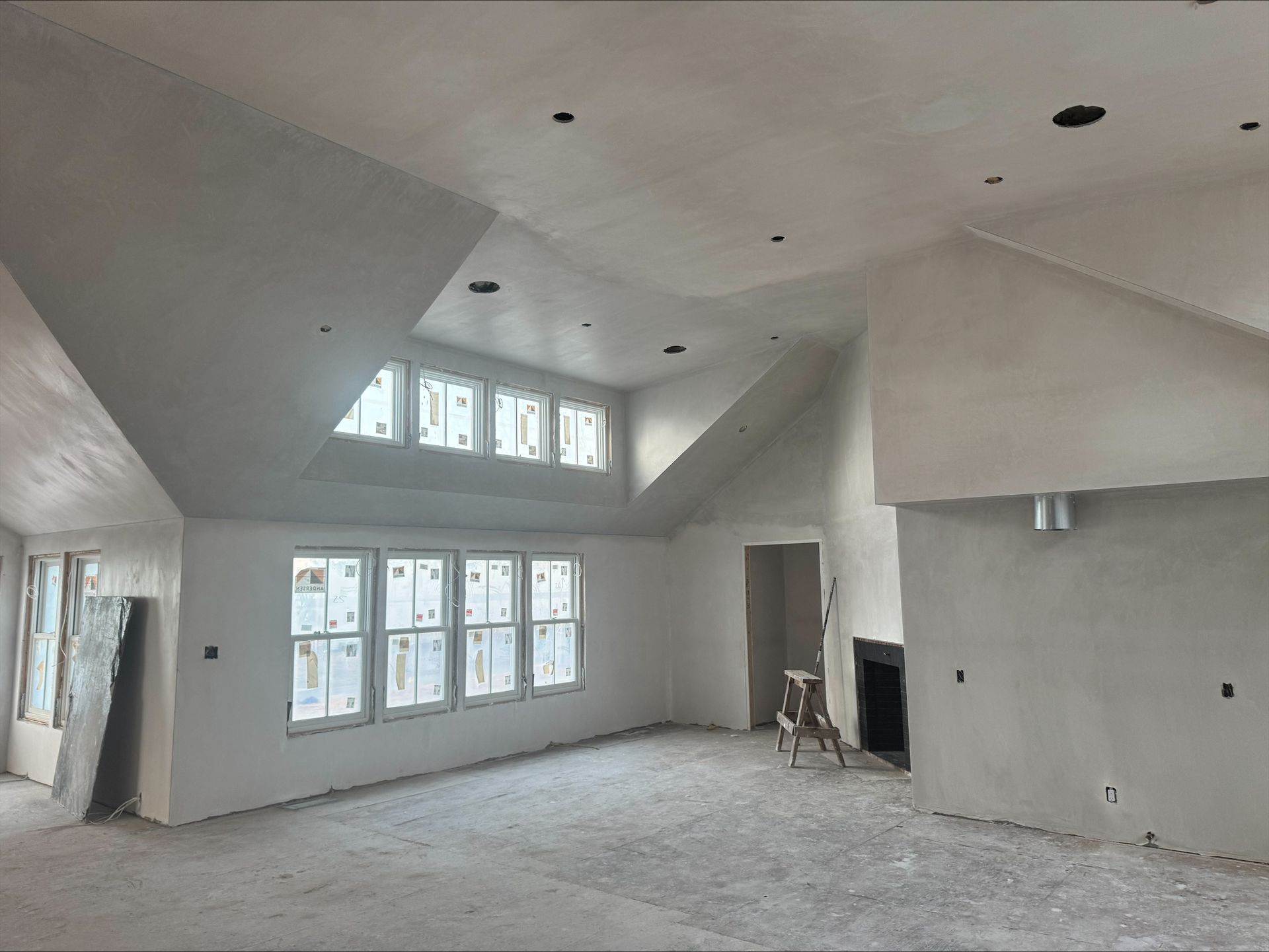 Empty unfinished living room with vaulted ceilings, large windows, and exposed drywall floor.