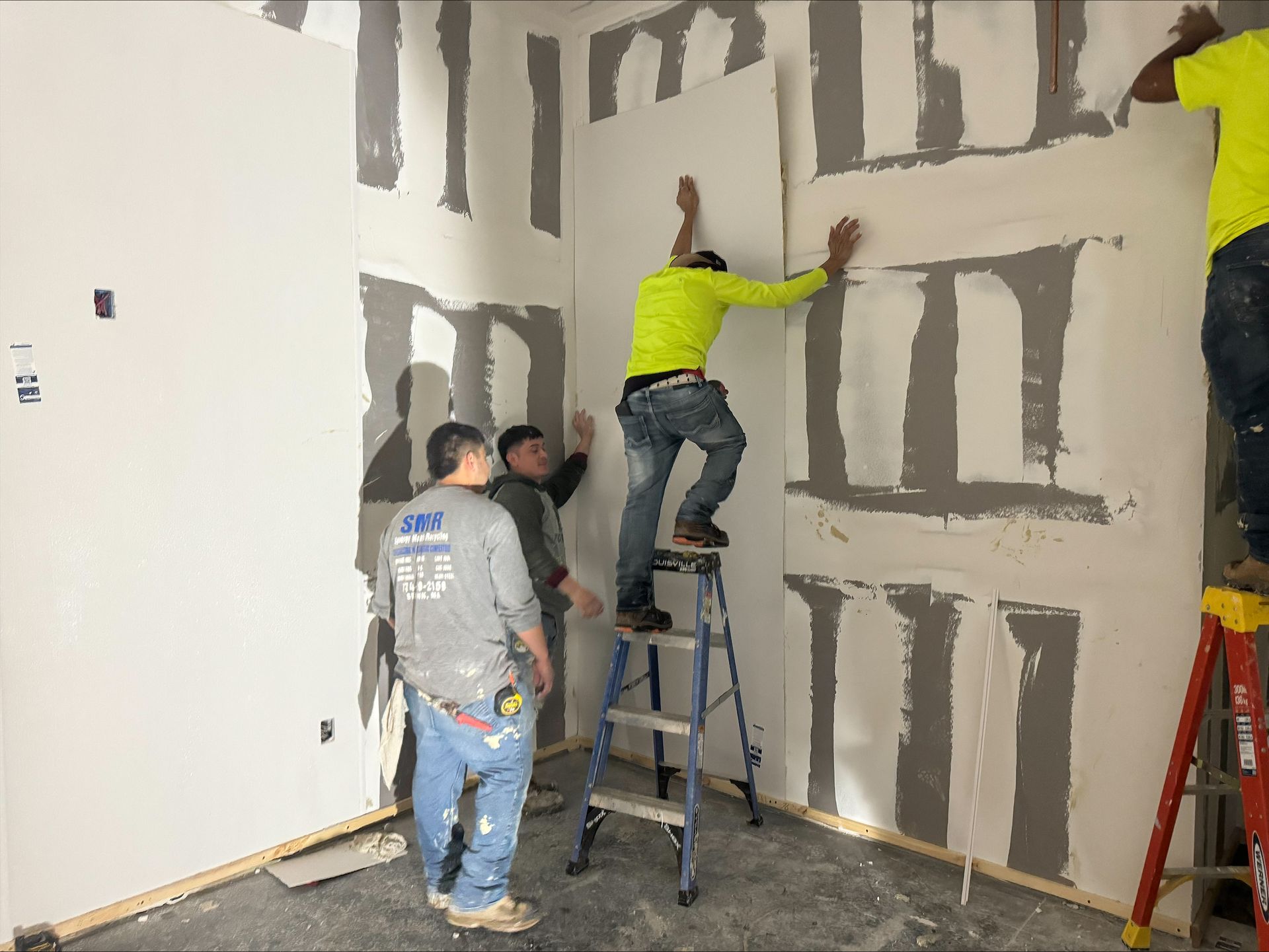 Workers painting a wall with gray block outlines; one on a ladder, two standing nearby.