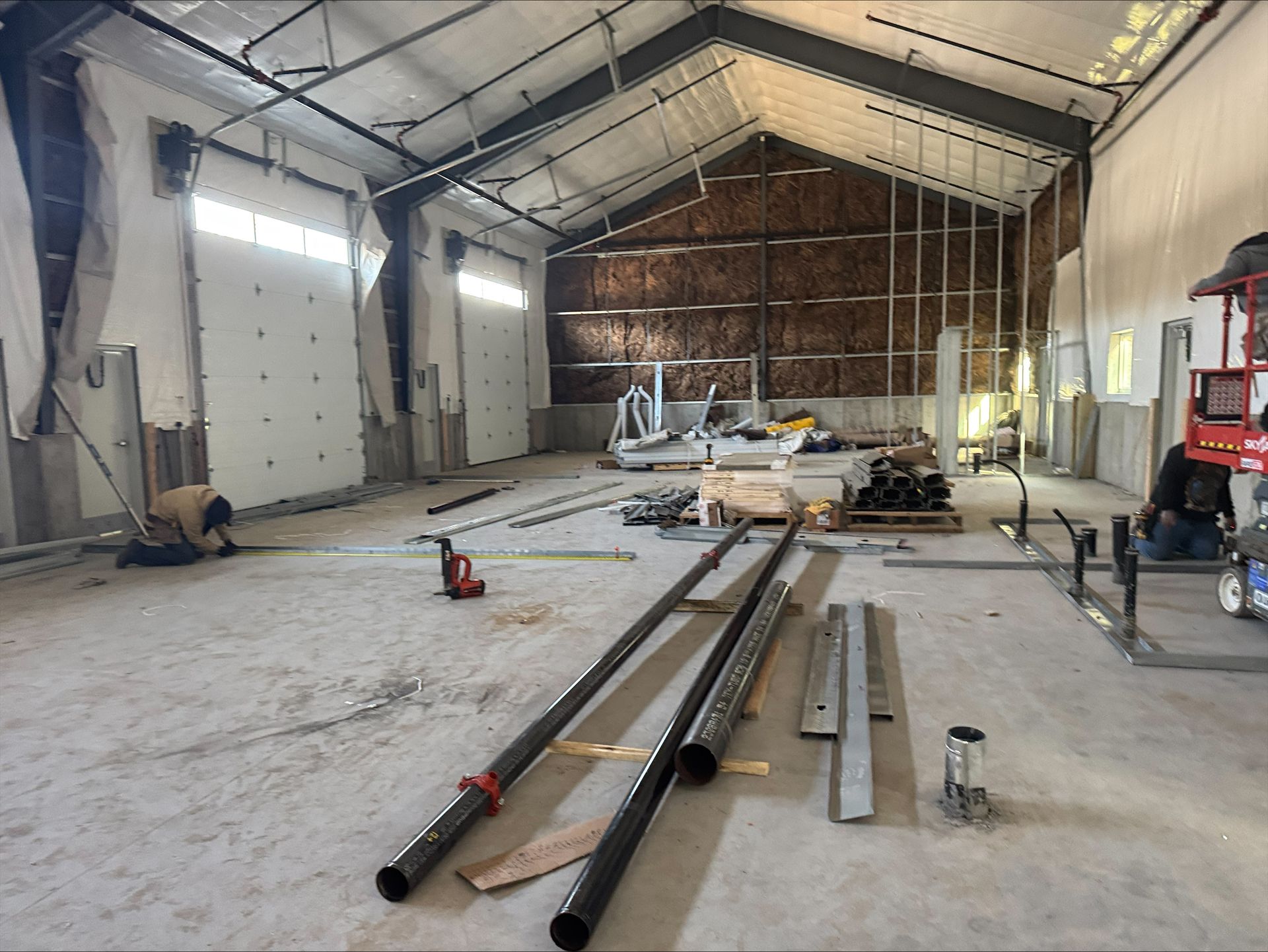 Inside a large industrial workshop, long metal beams and tools are laid out on a dusty concrete floor.