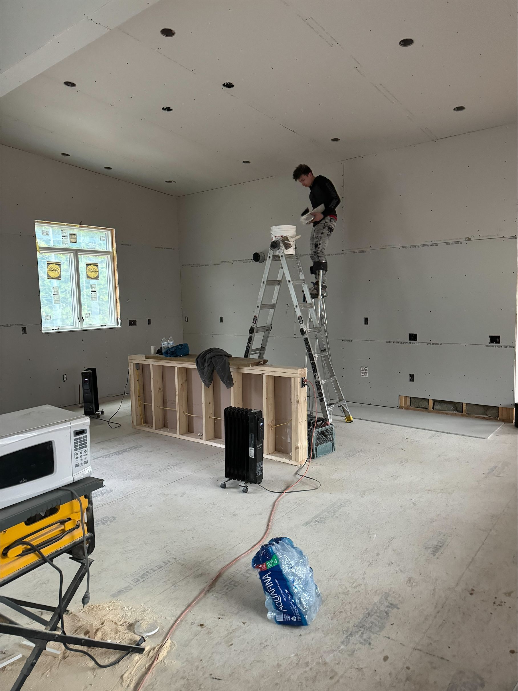 Interior room under renovation with a worker on a ladder over exposed wall framing and tools on the floor