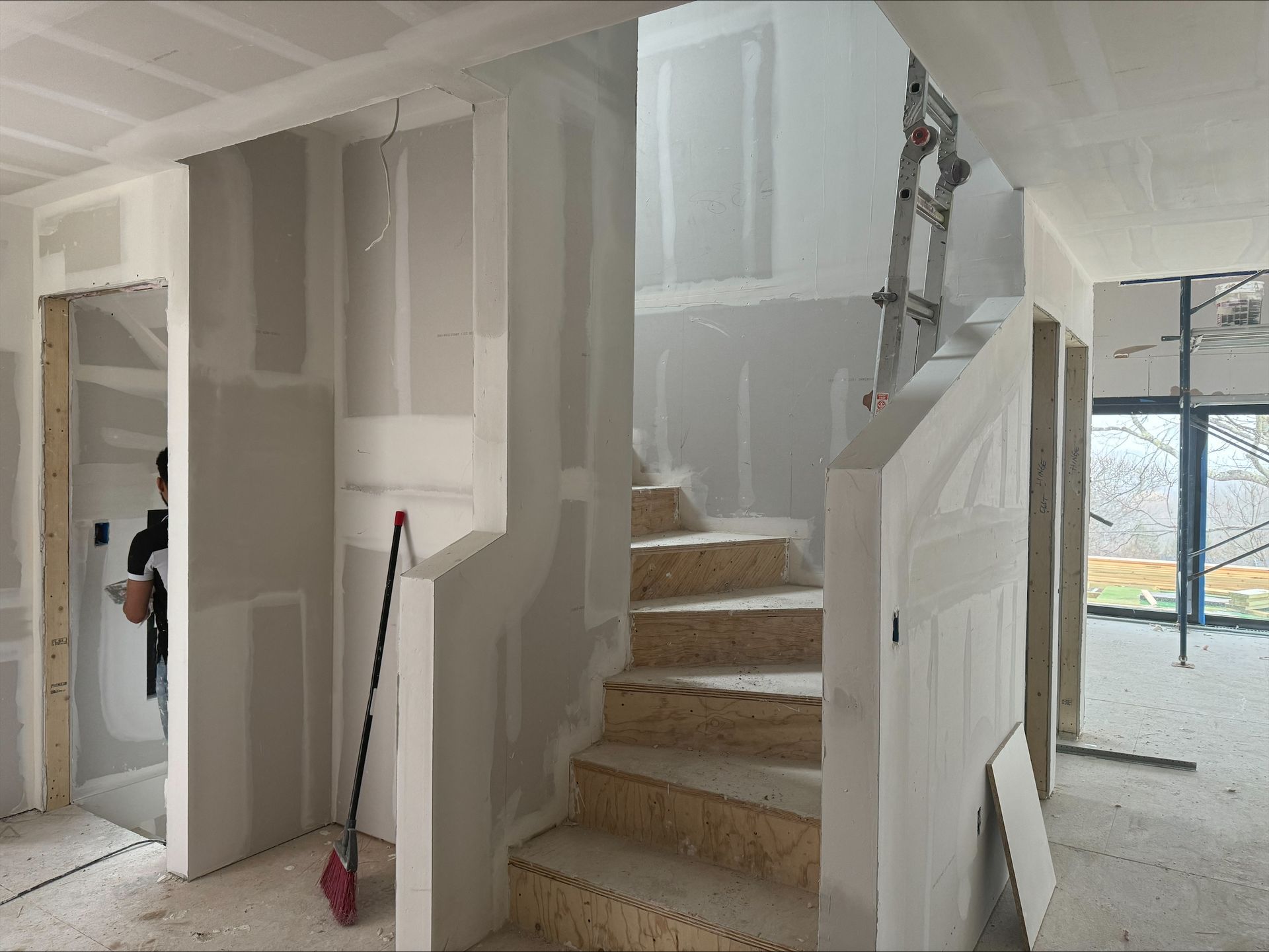 Partially finished staircase in an interior under construction, with drywall, ladder, and broom visible
