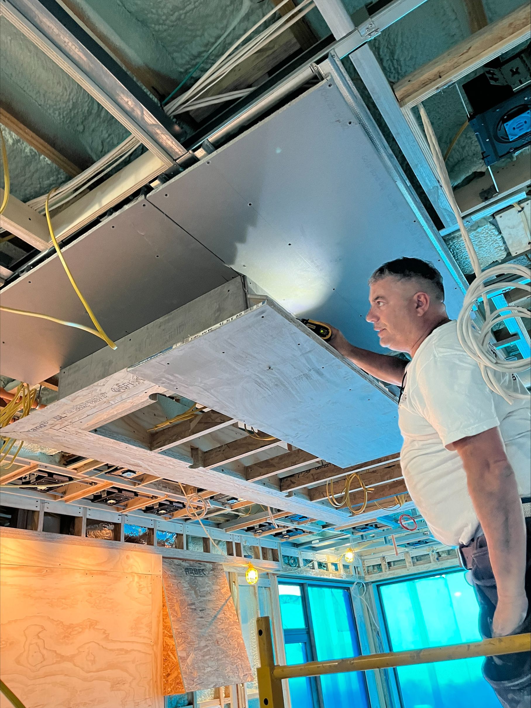 Man in white shirt inspecting ceiling panel in an unfinished room with exposed framing and blue windows