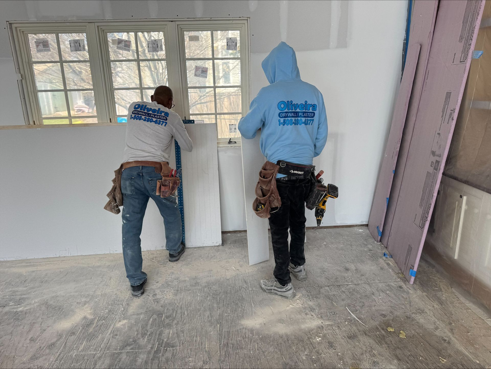 Two workers moving white panels in a room under construction, with dust on the floor and windows behind them