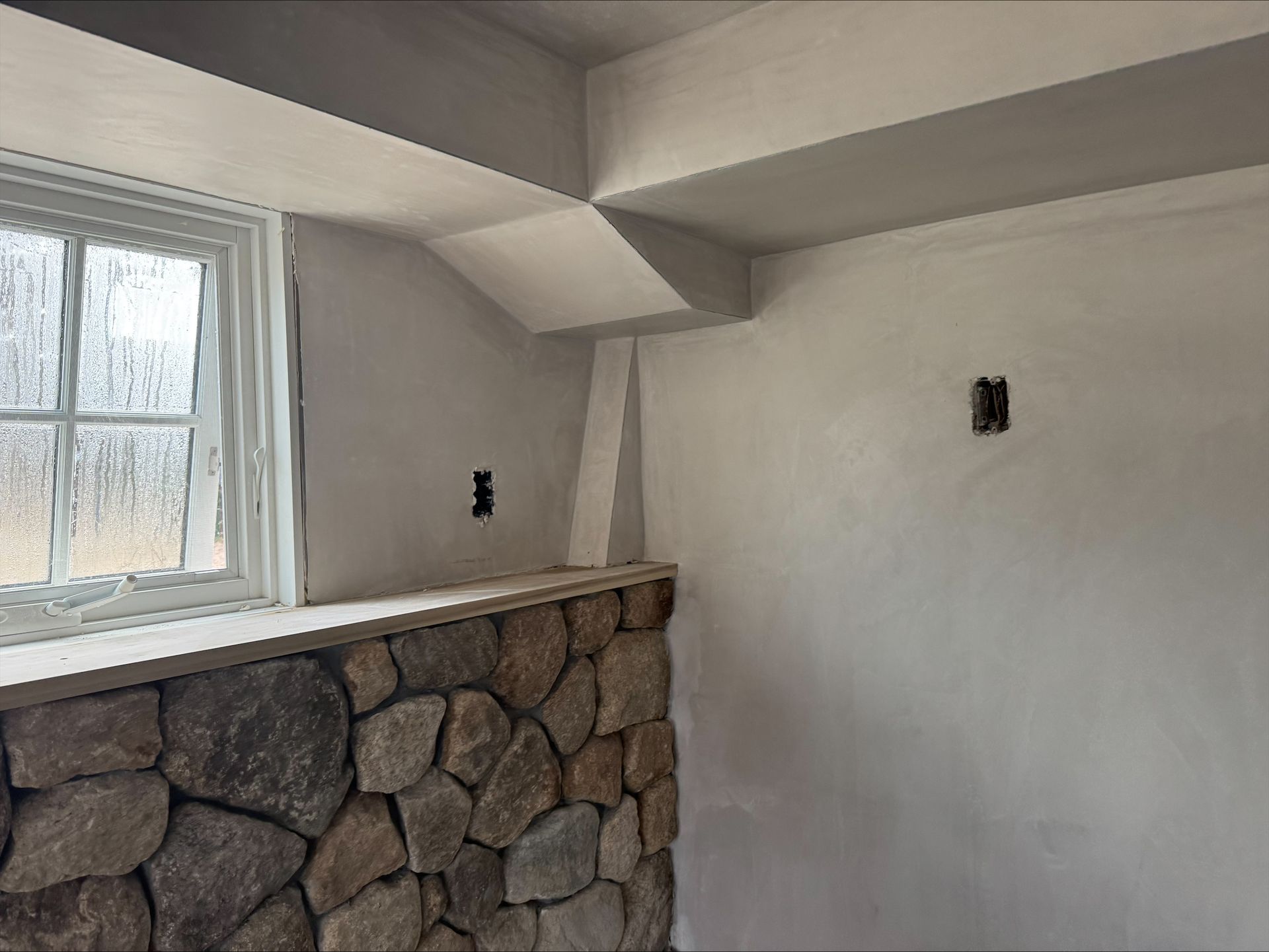 Small room with a stone half-wall, white walls and ceiling, and a window letting in daylight.