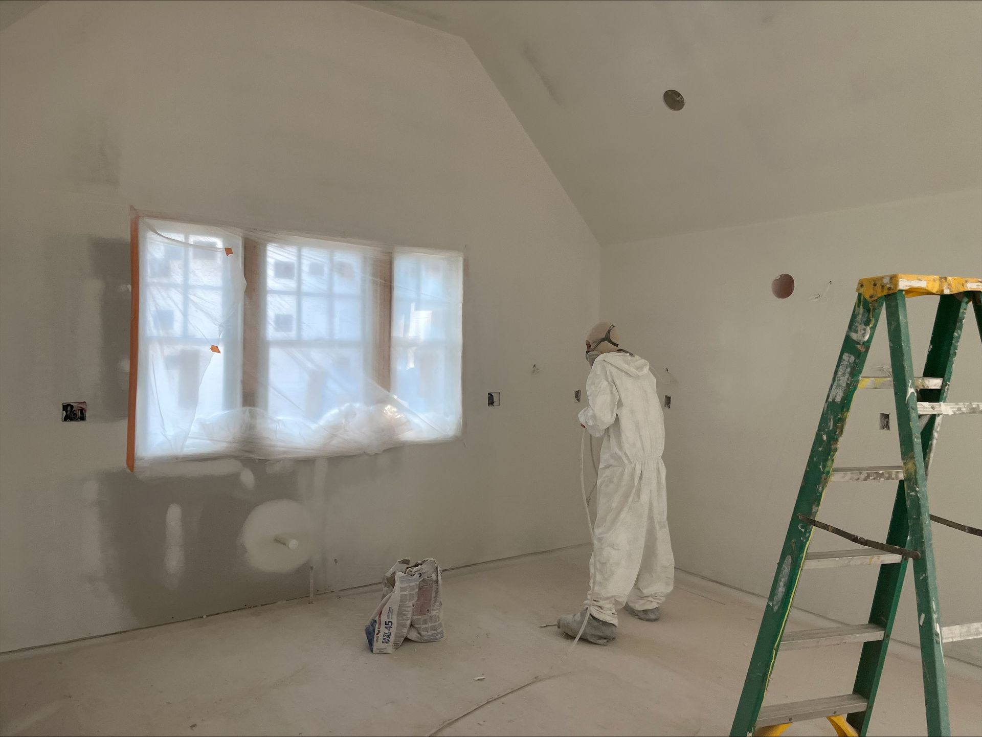 Worker in white coveralls painting a room’s white walls, with a green ladder nearby.