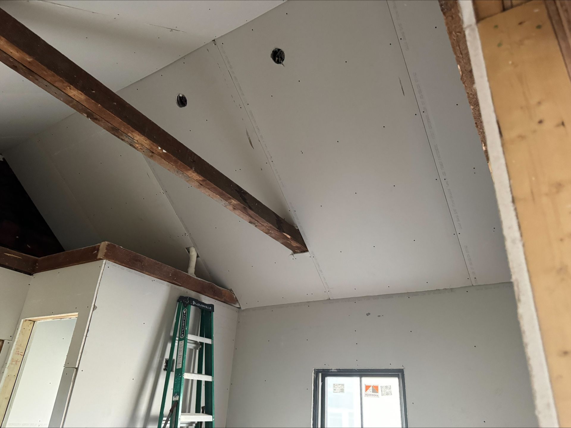 Interior room under construction with sloped drywall ceiling, exposed wooden beams, ladder, and window