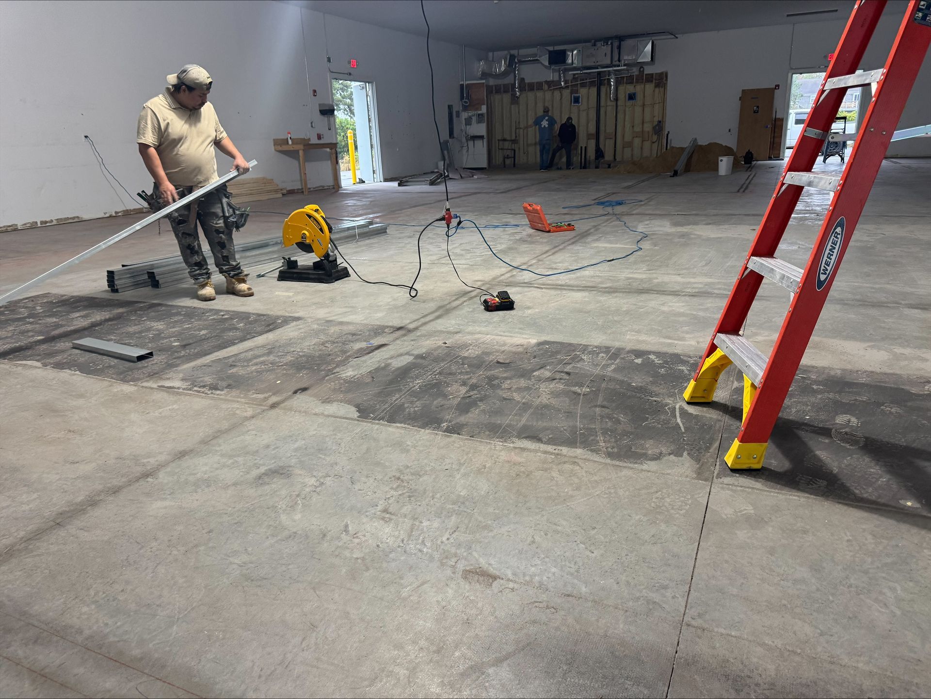 Worker using a floor saw inside a garage, with tools and a red ladder nearby.
