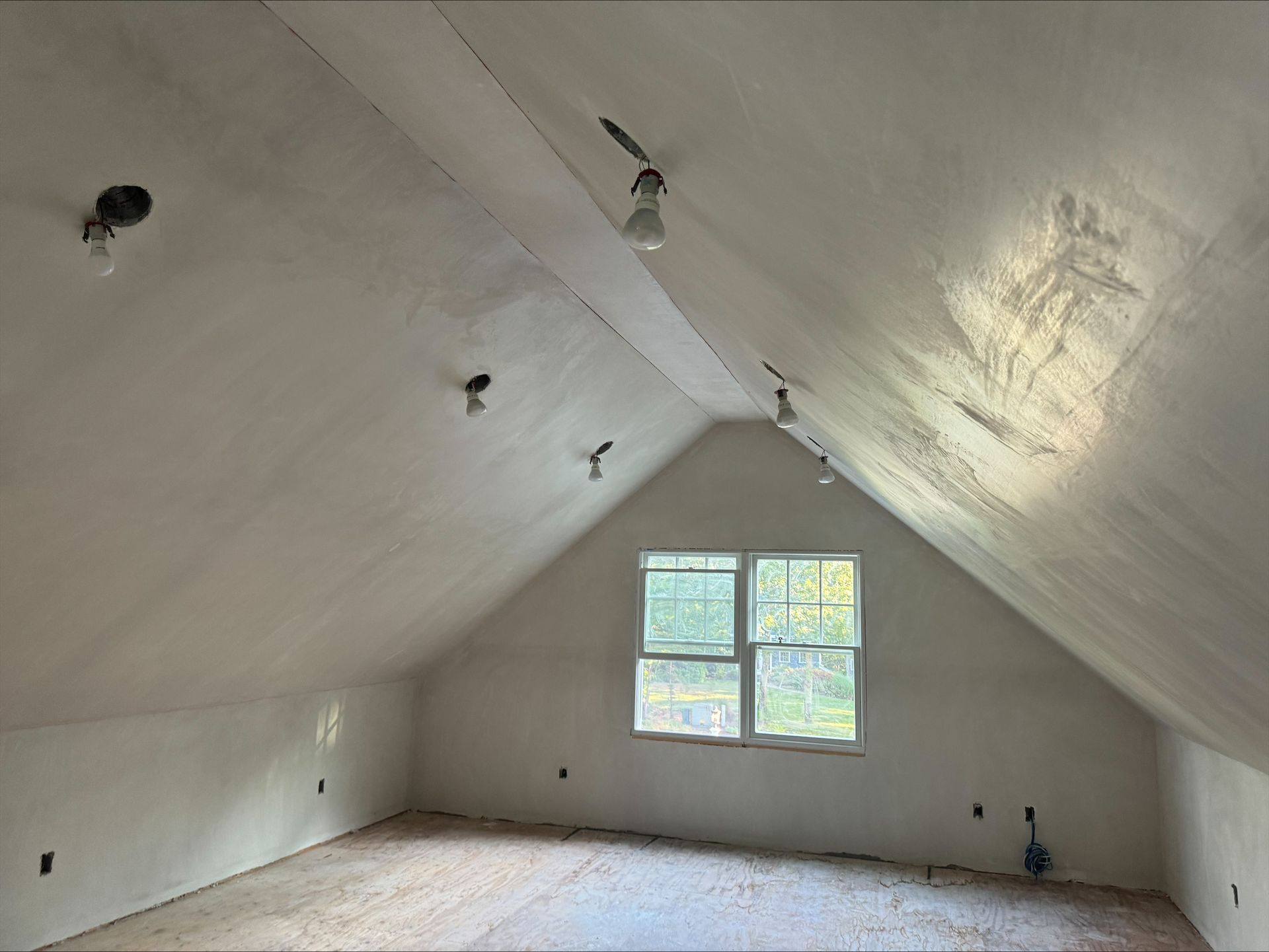 Empty attic room with sloped white walls, exposed lights, and a small window