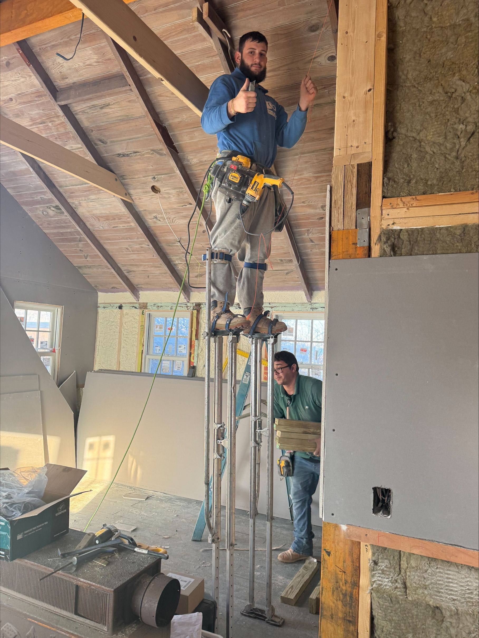 Worker standing on a tall scaffold inside an unfinished room with exposed beams and insulation.