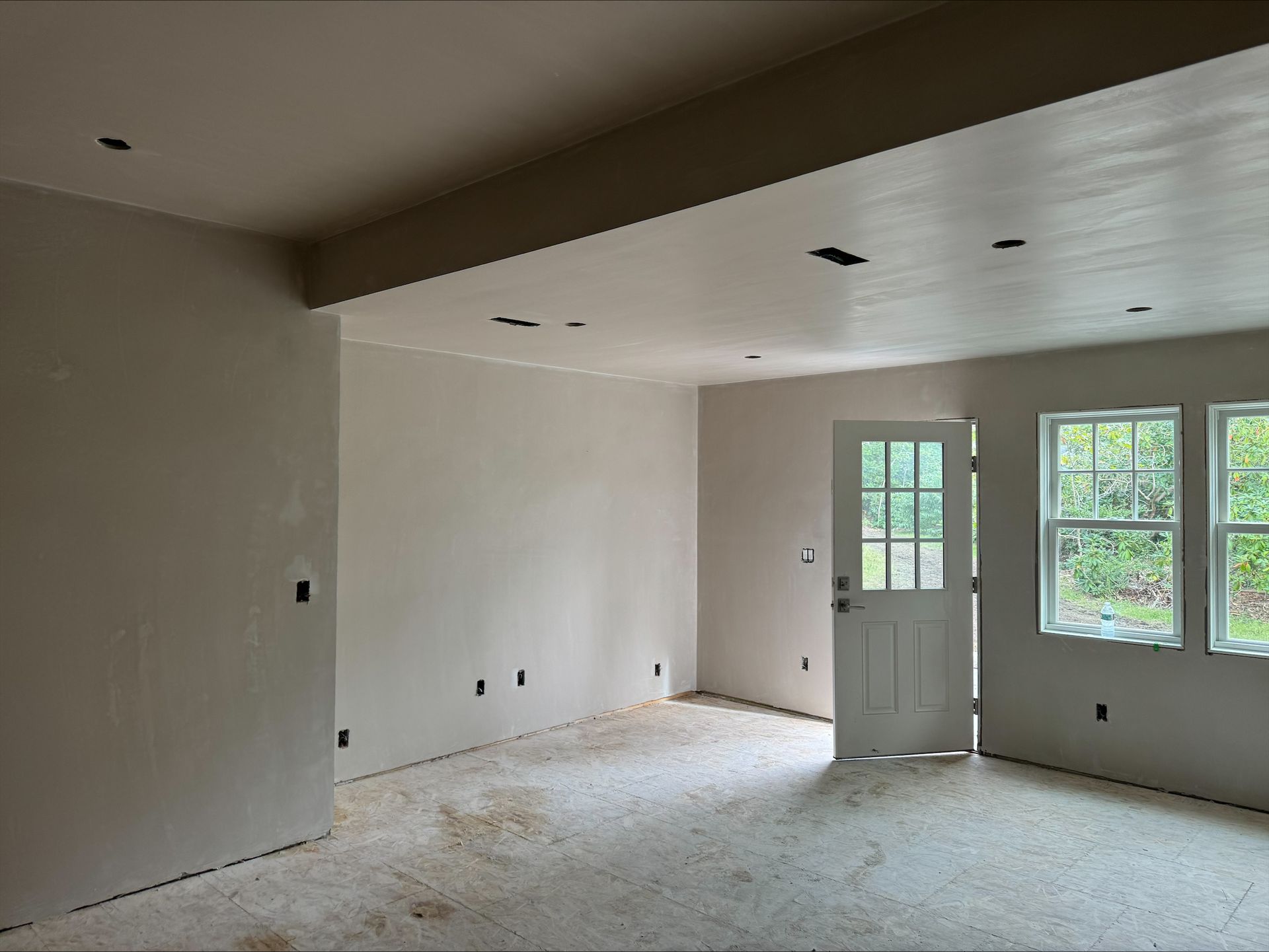 Empty unfinished room with white walls, recessed ceiling lights, windows, and a glass door.