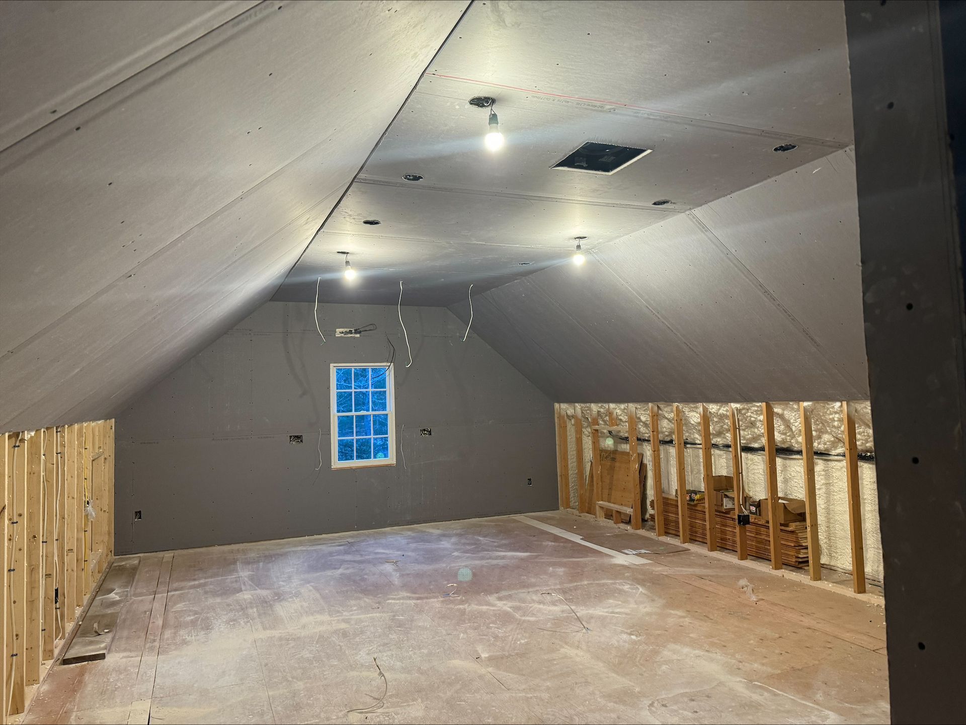 Attic room under renovation with exposed framing, sloped ceiling, and a small window at the far end