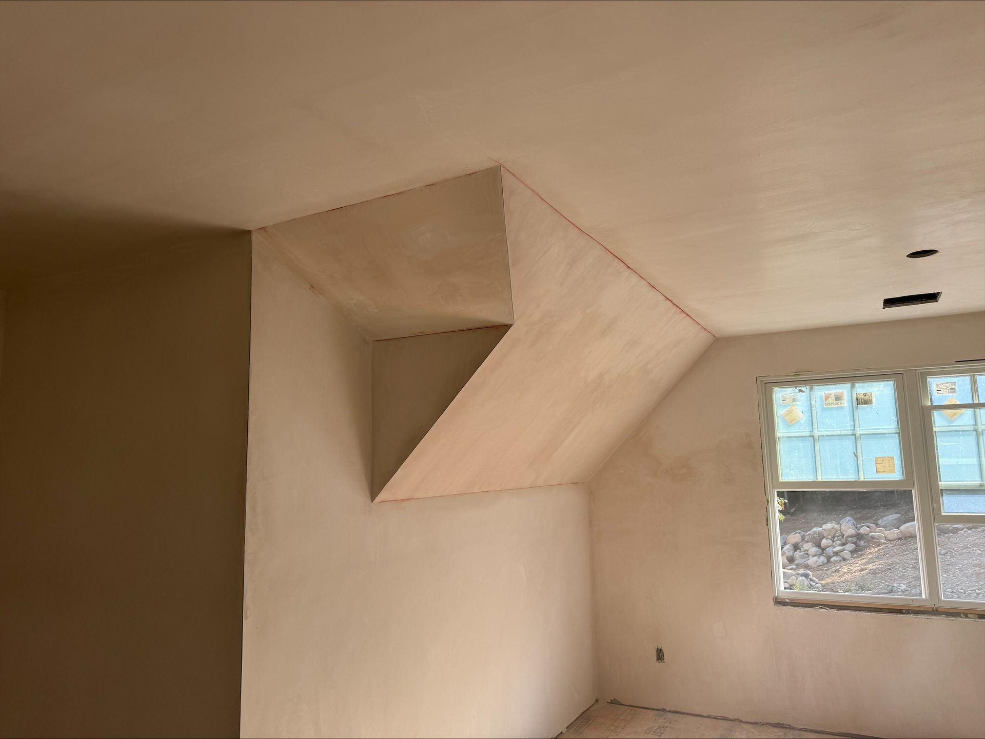 Empty beige room with a large geometric opening near the ceiling and a window on the right.