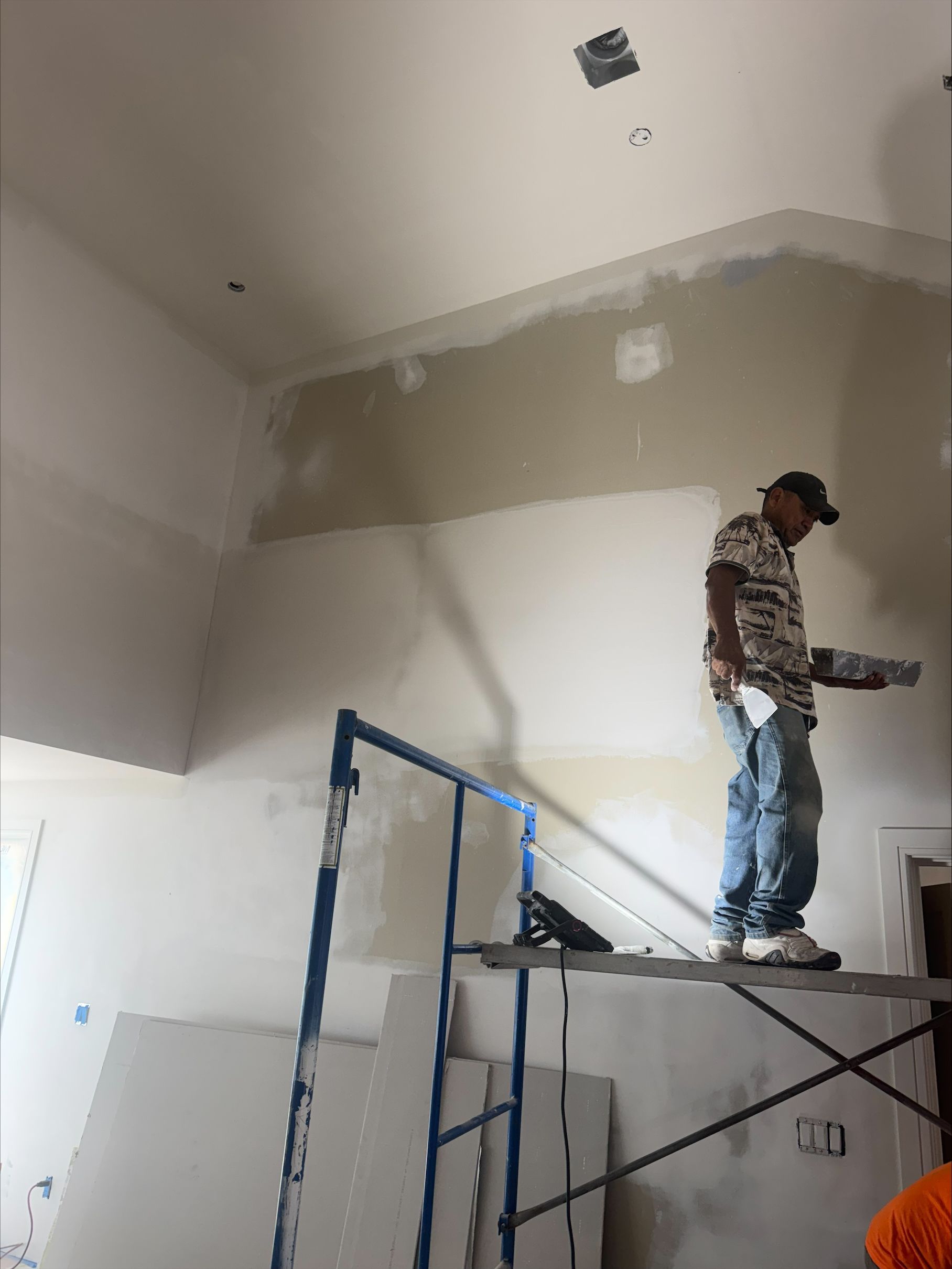 Worker standing on scaffolding, painting a high wall and ceiling in a room under renovation.