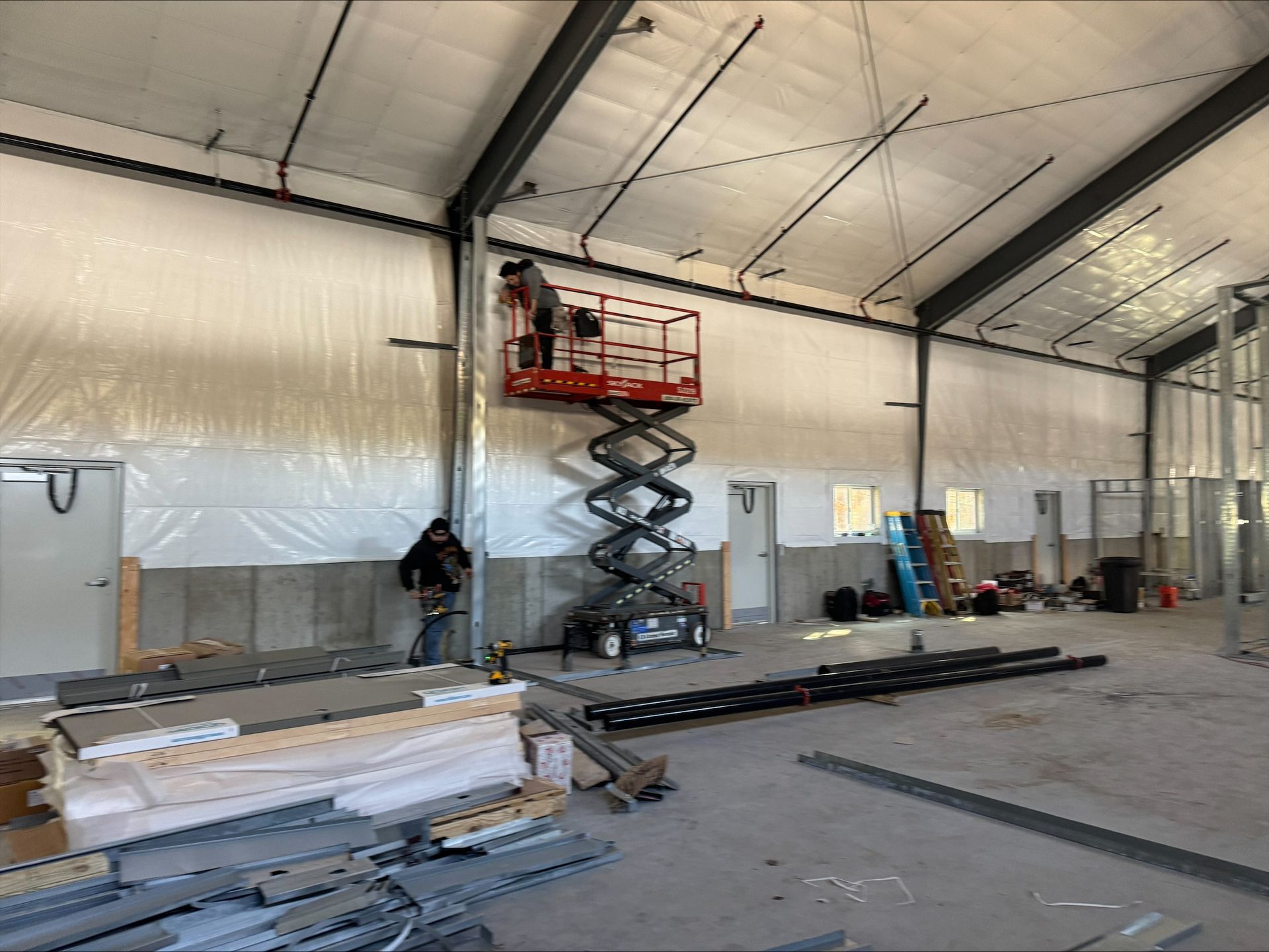 Interior of a warehouse under construction with a scissor lift, stacked materials, and plastic-covered walls