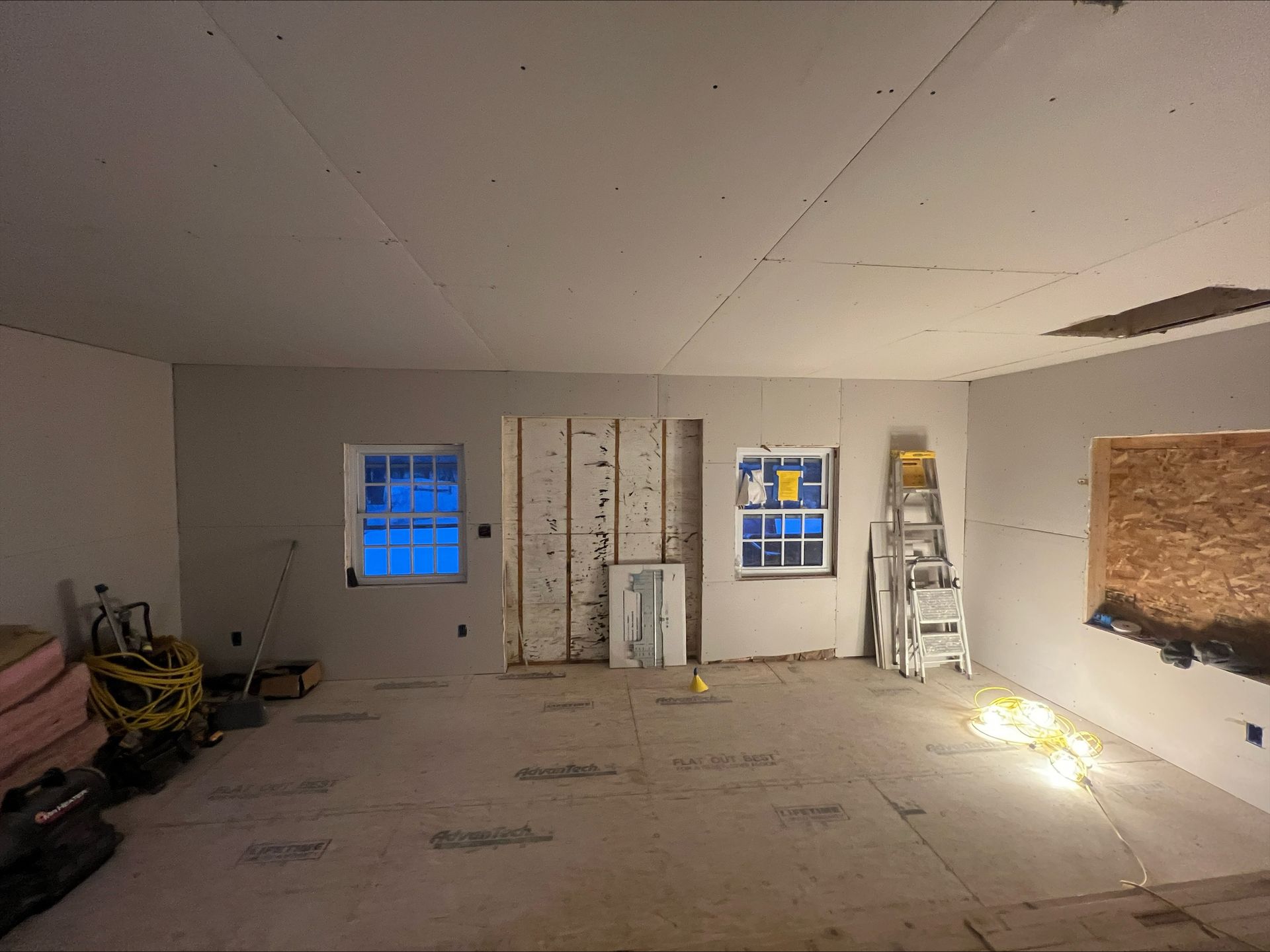 Empty unfinished room with drywall, two windows, ladder, and construction materials on the floor.