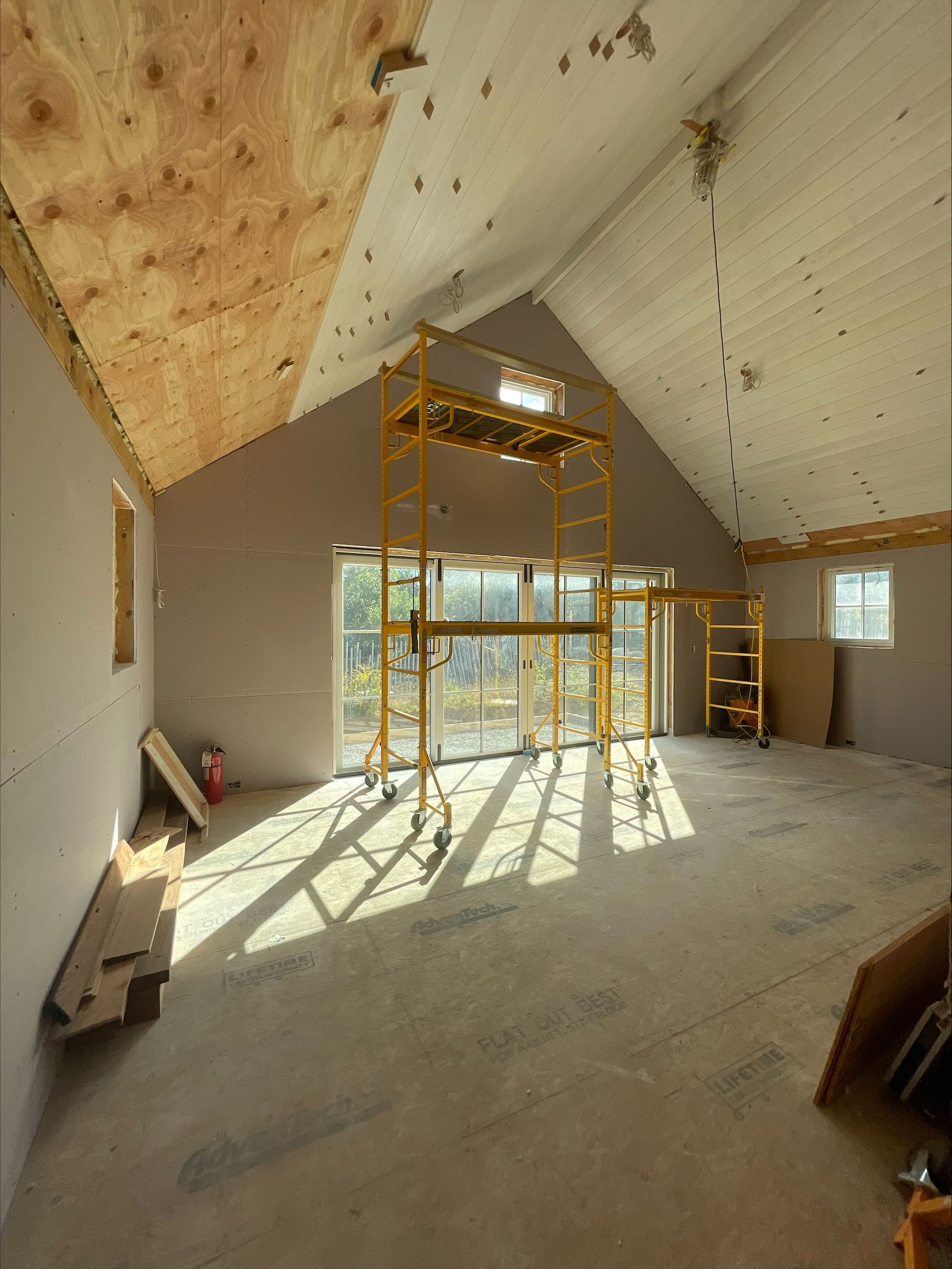Interior of a high-ceiling room under construction with scaffolding, exposed insulation, and sunlight through large windows