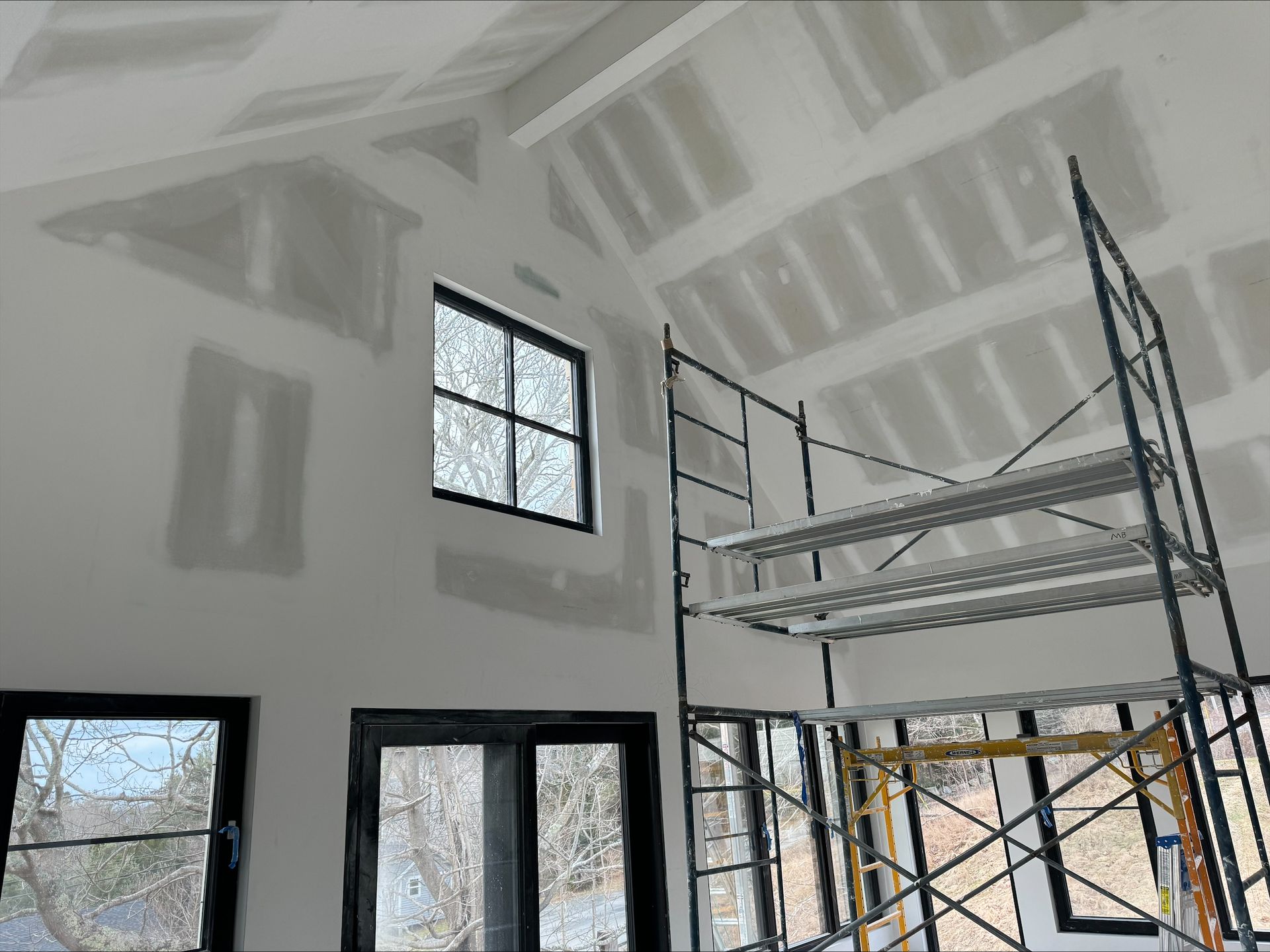 Interior of a house under construction with drywall, scaffolding, and large windows.