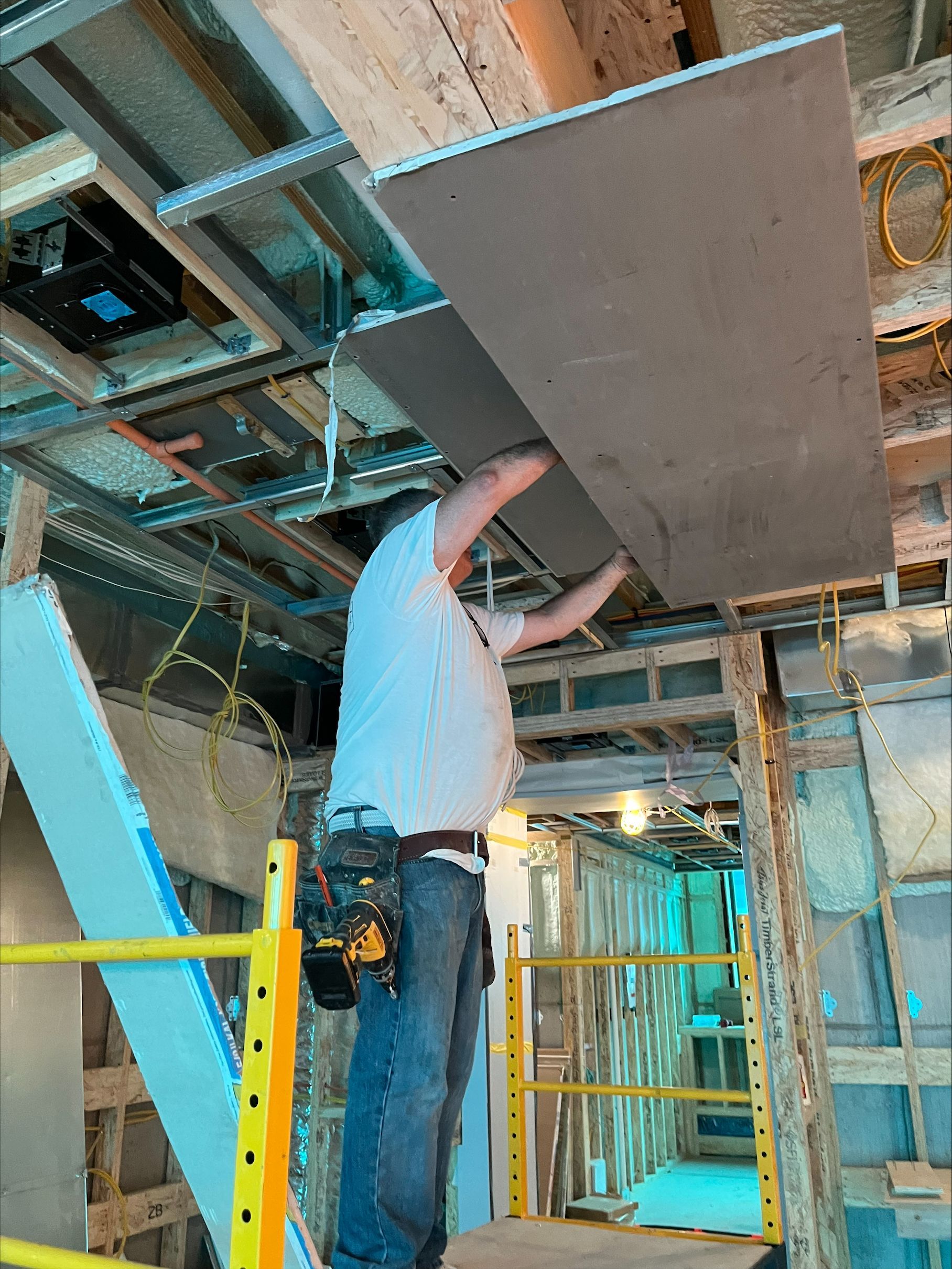Worker installing a ceiling panel on a ladder in an unfinished room with exposed beams