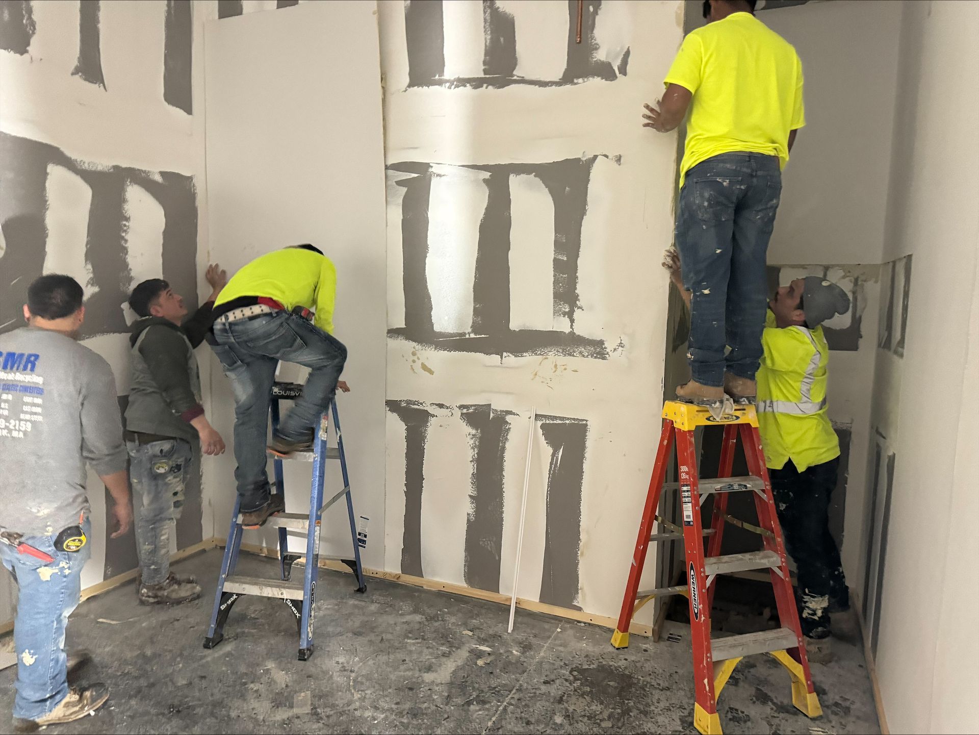 Workers plastering a wall indoors, with ladders and buckets at a construction site