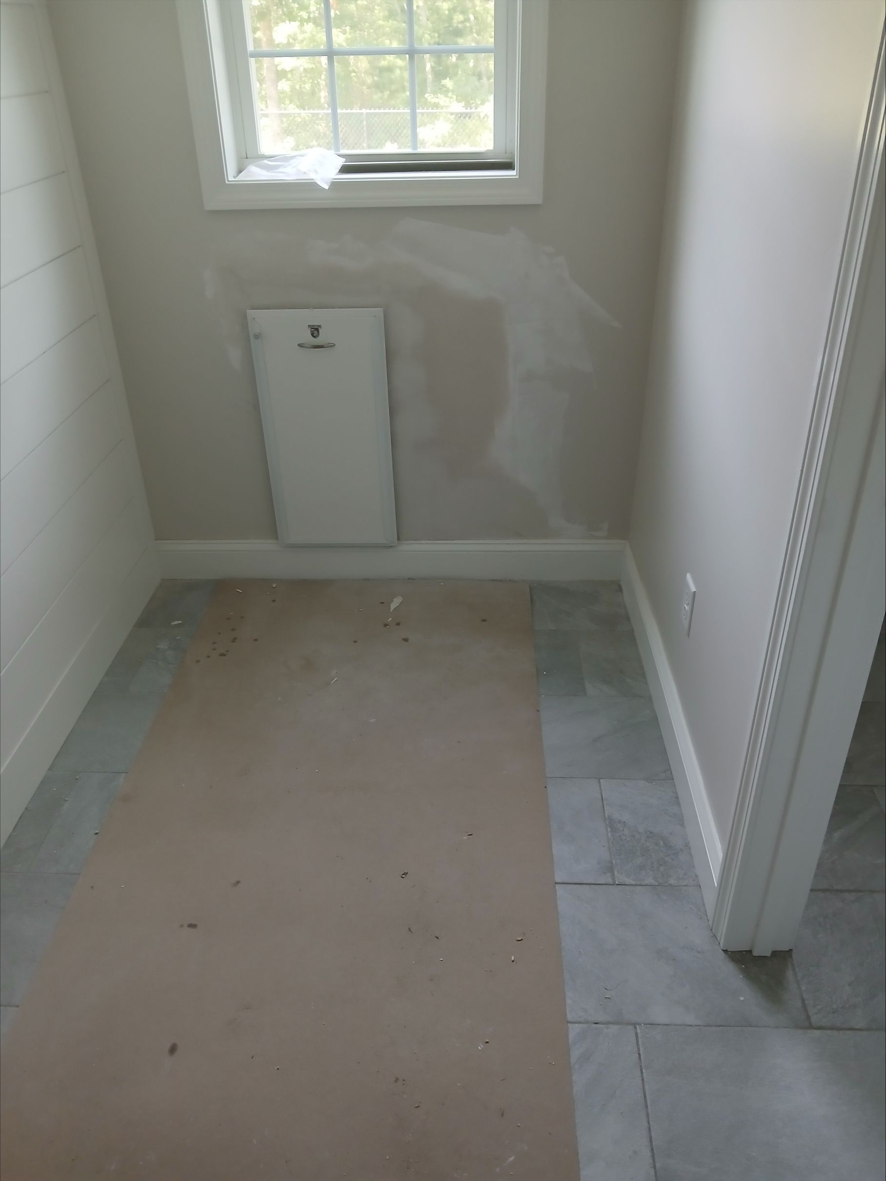 Small unfinished bathroom with window, toilet access panel, and partially removed floor tiles.