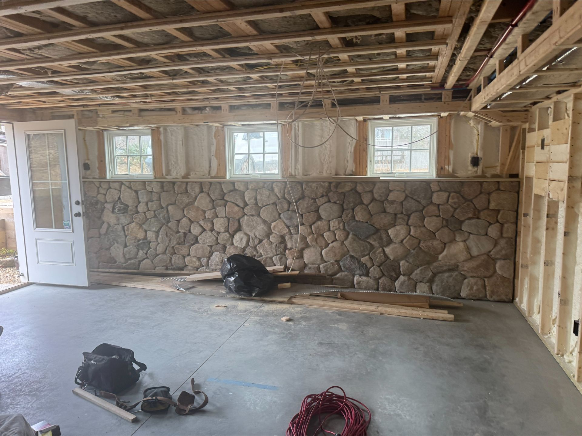 Unfinished garage with exposed ceiling, stone half-wall, open door, and tools on the concrete floor.