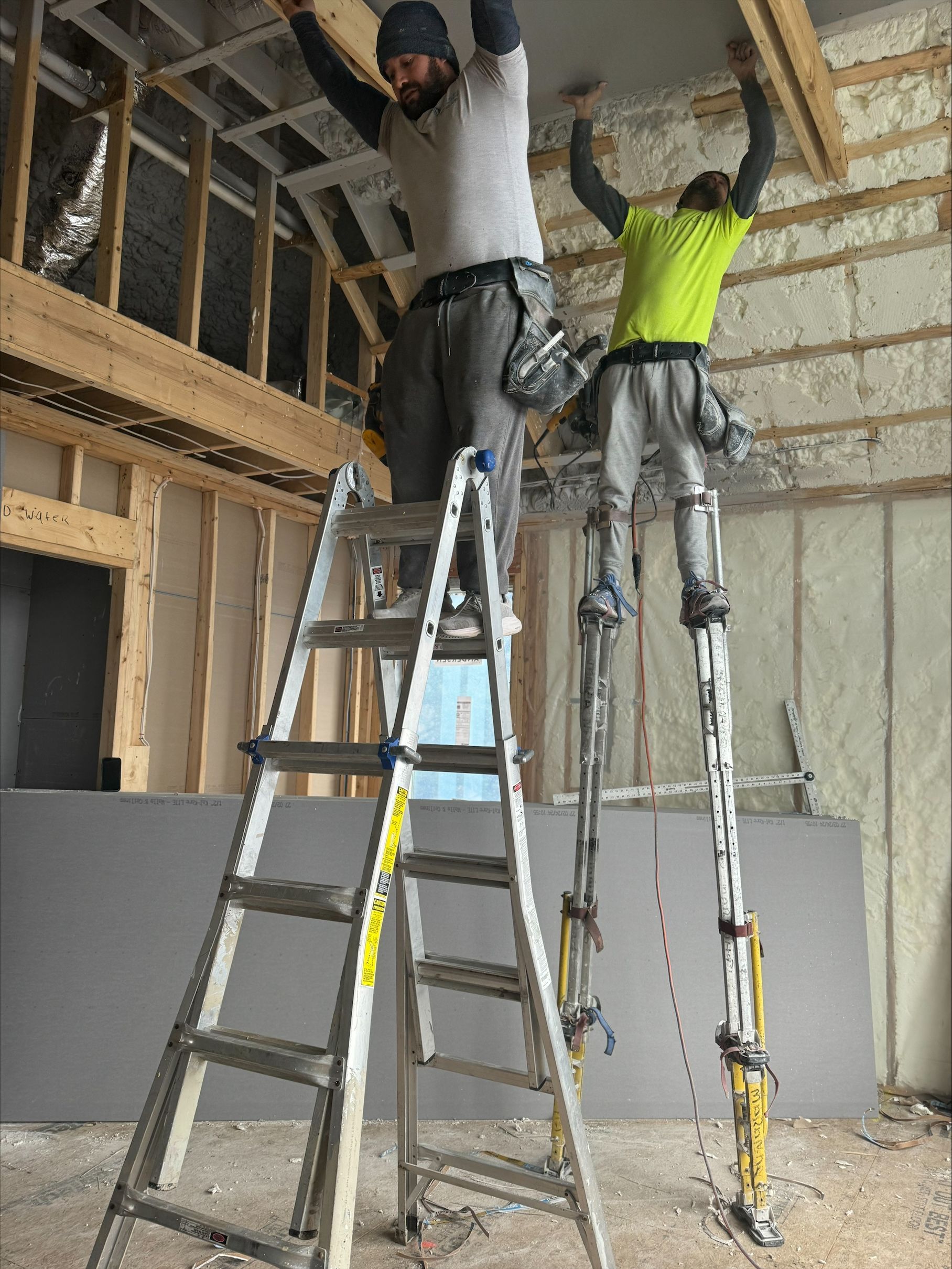 Two workers in a construction site standing on ladders, reaching up to install ceiling framing.