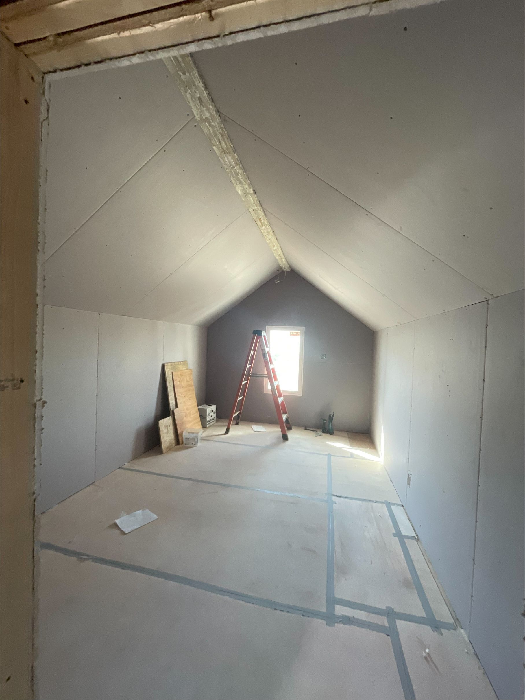 Empty unfinished attic room with sloped white walls, ladder, and a small window at the far end