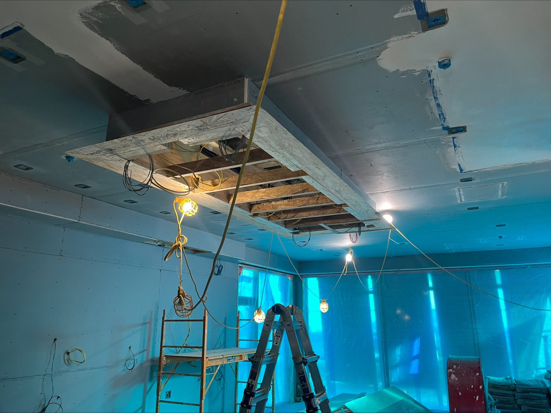 Interior under renovation with ladders and exposed ceiling panels in a blue-lit room
