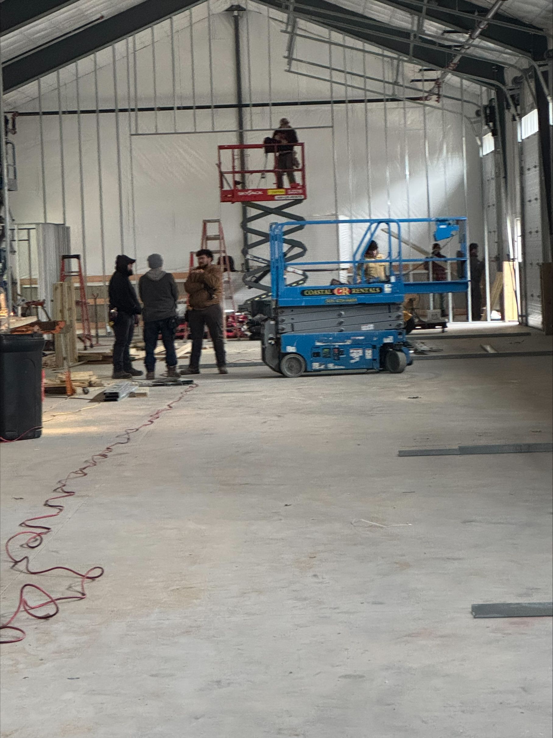 Workers and a red-and-blue lift inside an unfinished warehouse with a high metal roof