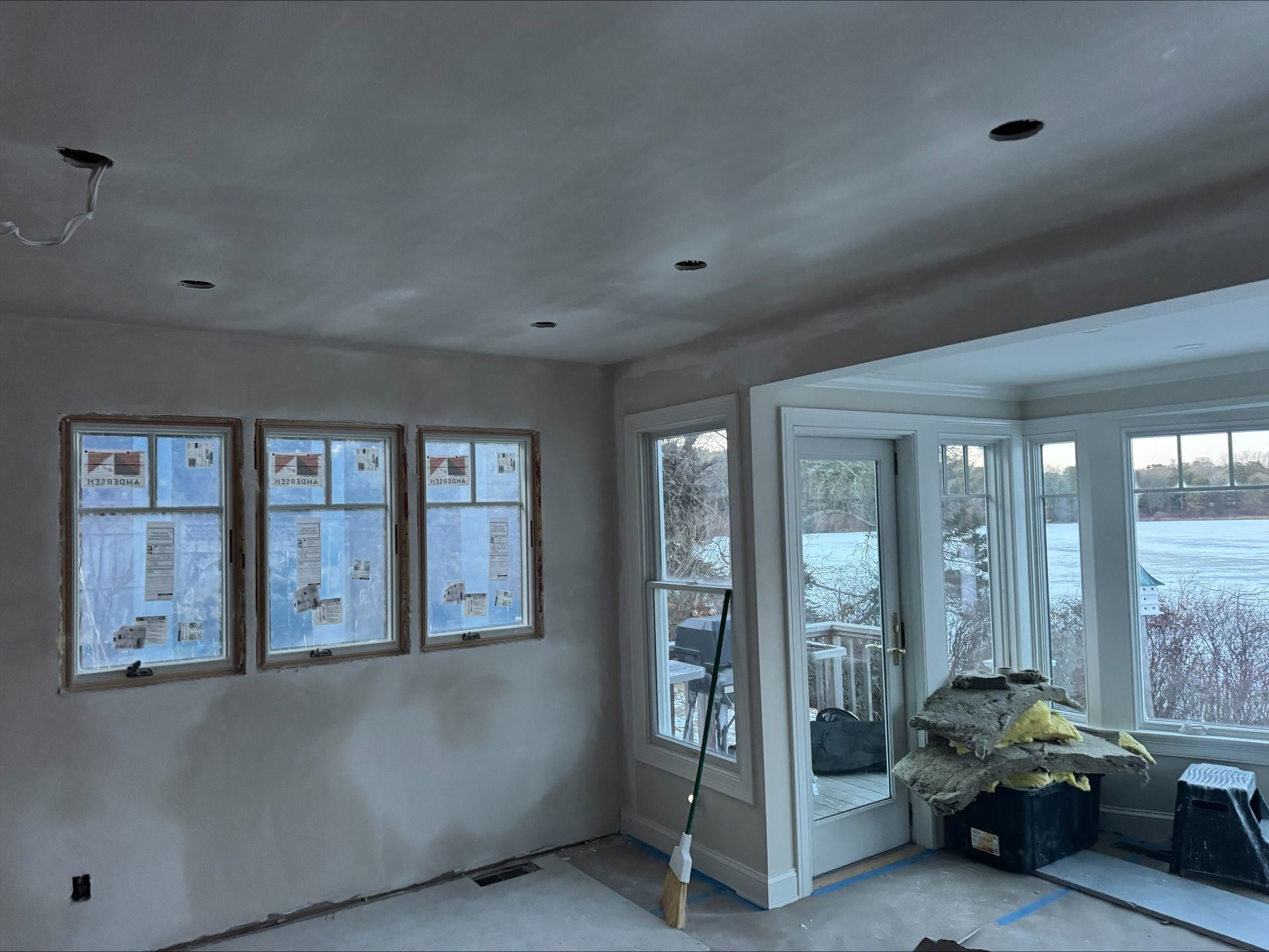 Unfinished room with white walls, exposed ceiling lights, windows, and a sliding glass door to a sunroom.