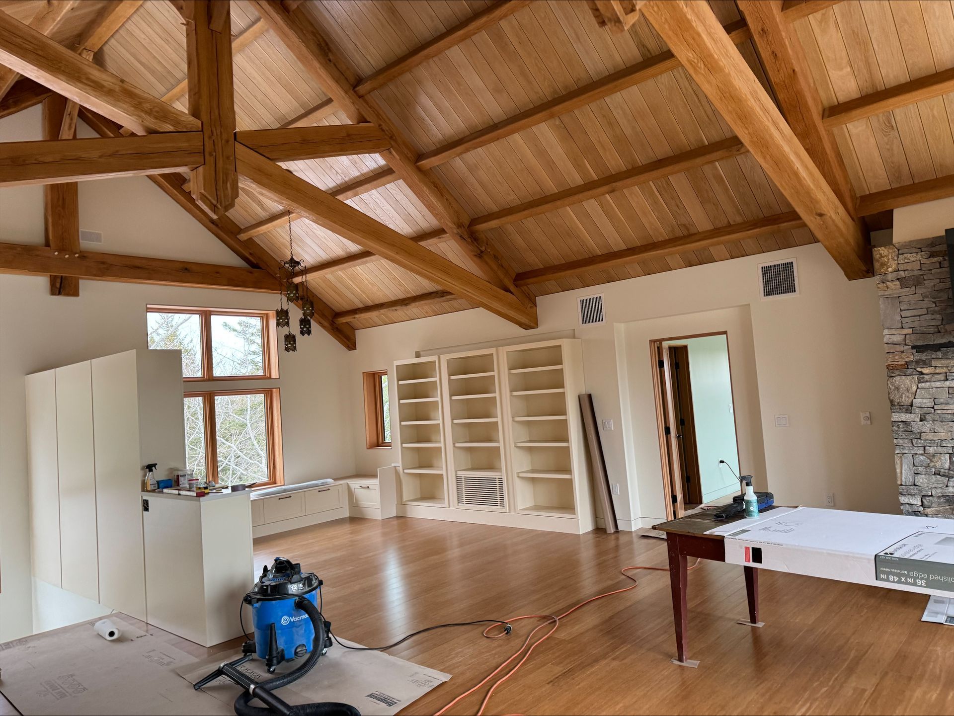 Wooden-beamed room with sloped ceiling, built-in shelves, table, and cleaning supplies on a polished floor