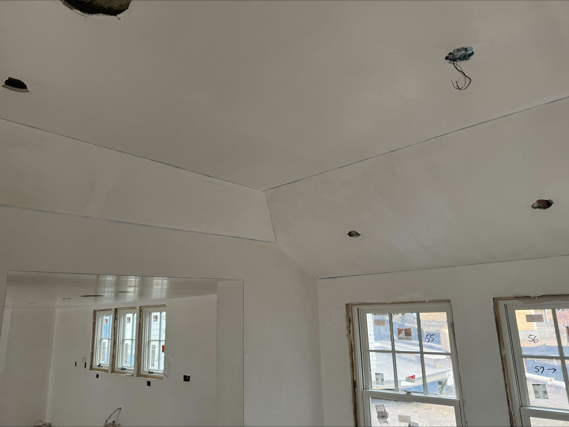 White unfinished room ceiling with recessed light openings and windows along the wall
