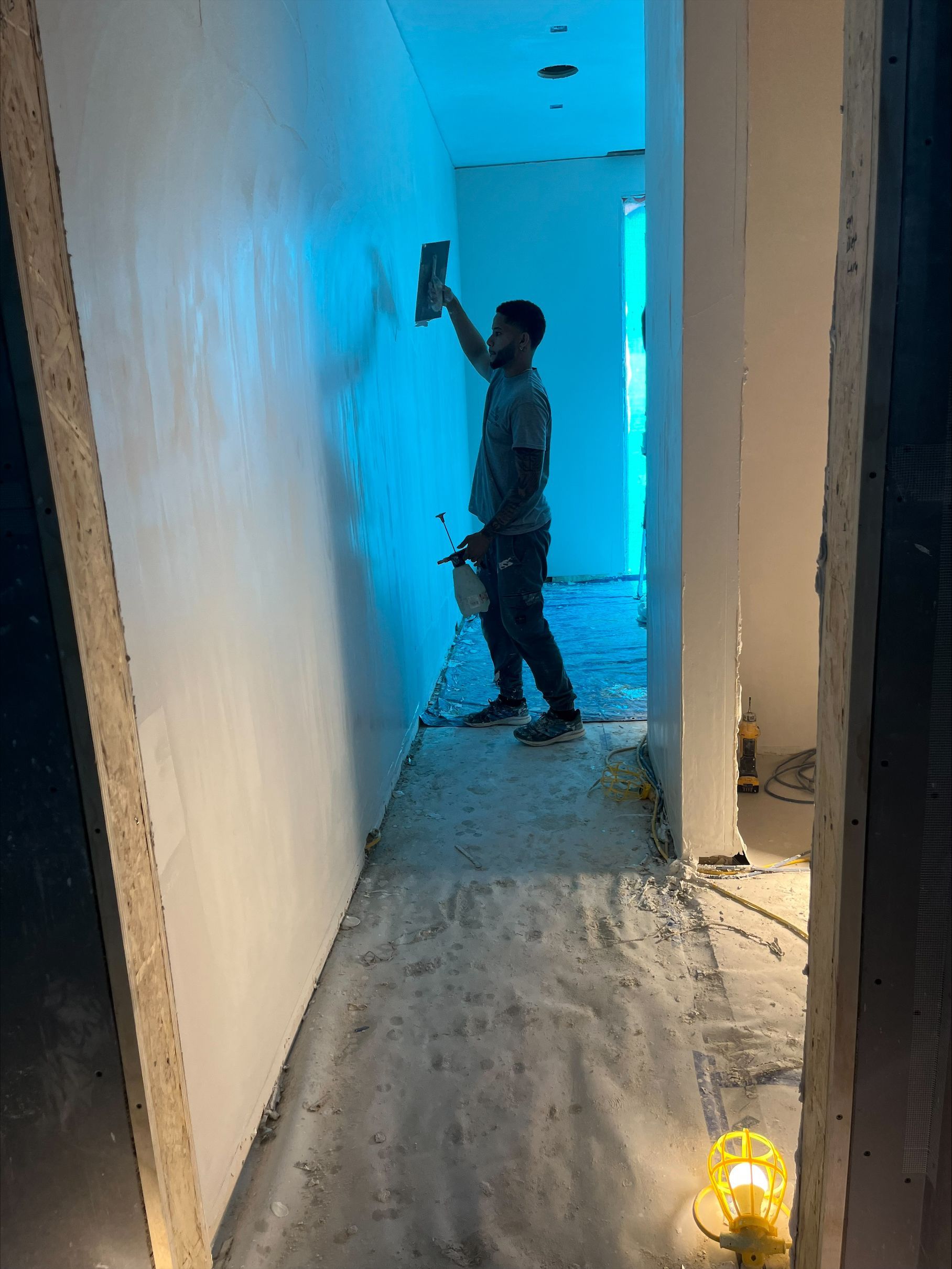 Worker painting a narrow hallway with blue walls and exposed flooring, yellow work light on the floor.