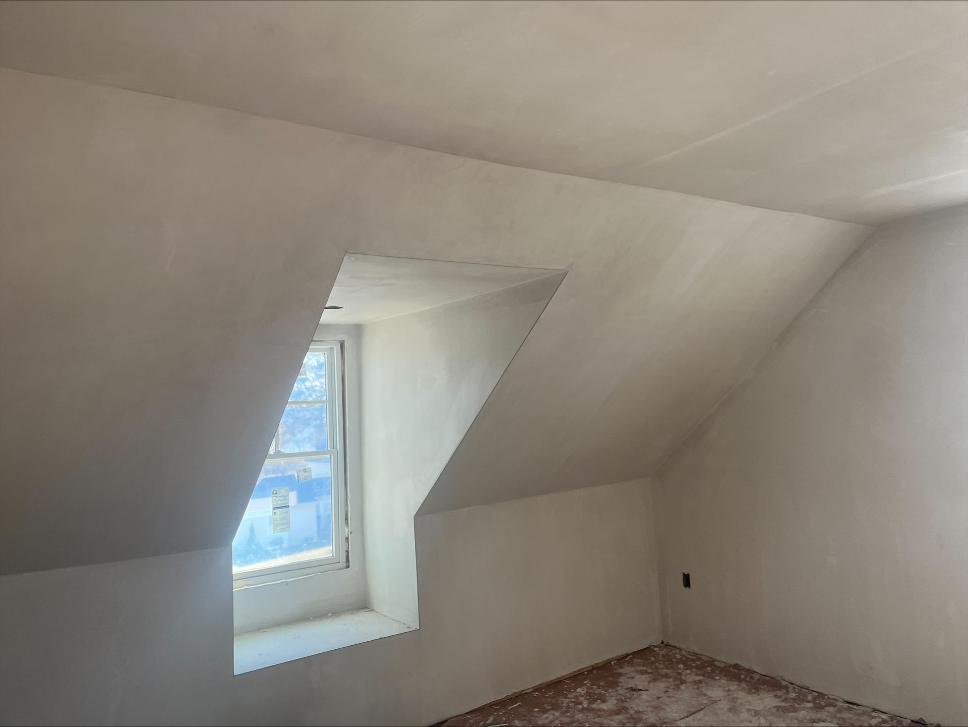 Empty attic room with slanted white walls, a small window, and bare floorboards.