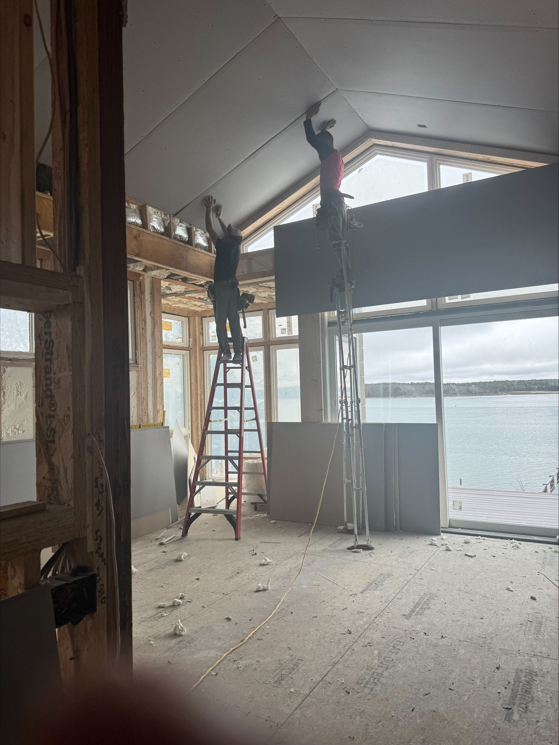 Two workers on ladders installing a ceiling in a bright, unfinished room overlooking water