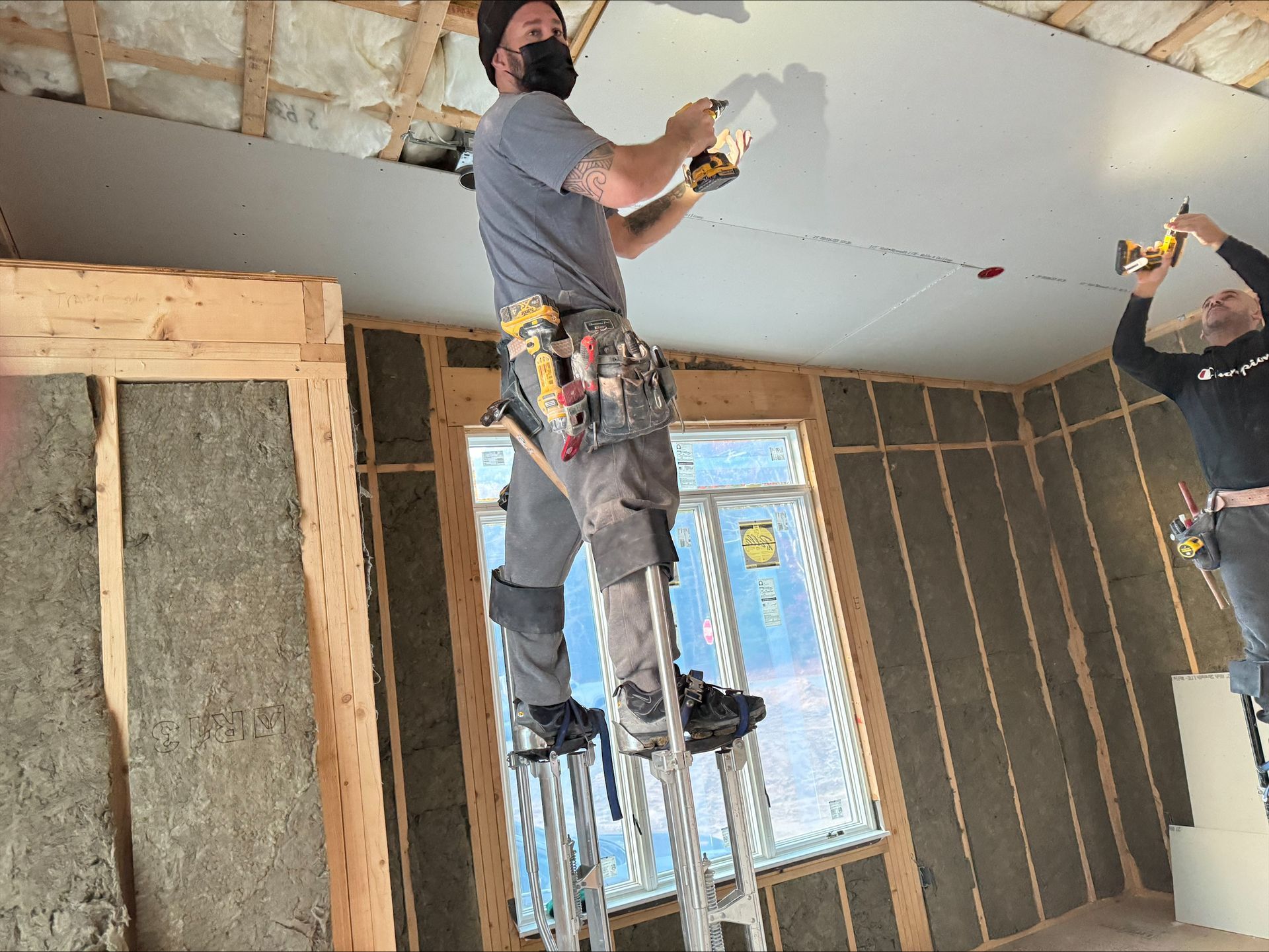 Workers installing drywall on a ceiling in a partially renovated room, using tools and standing on ladders.