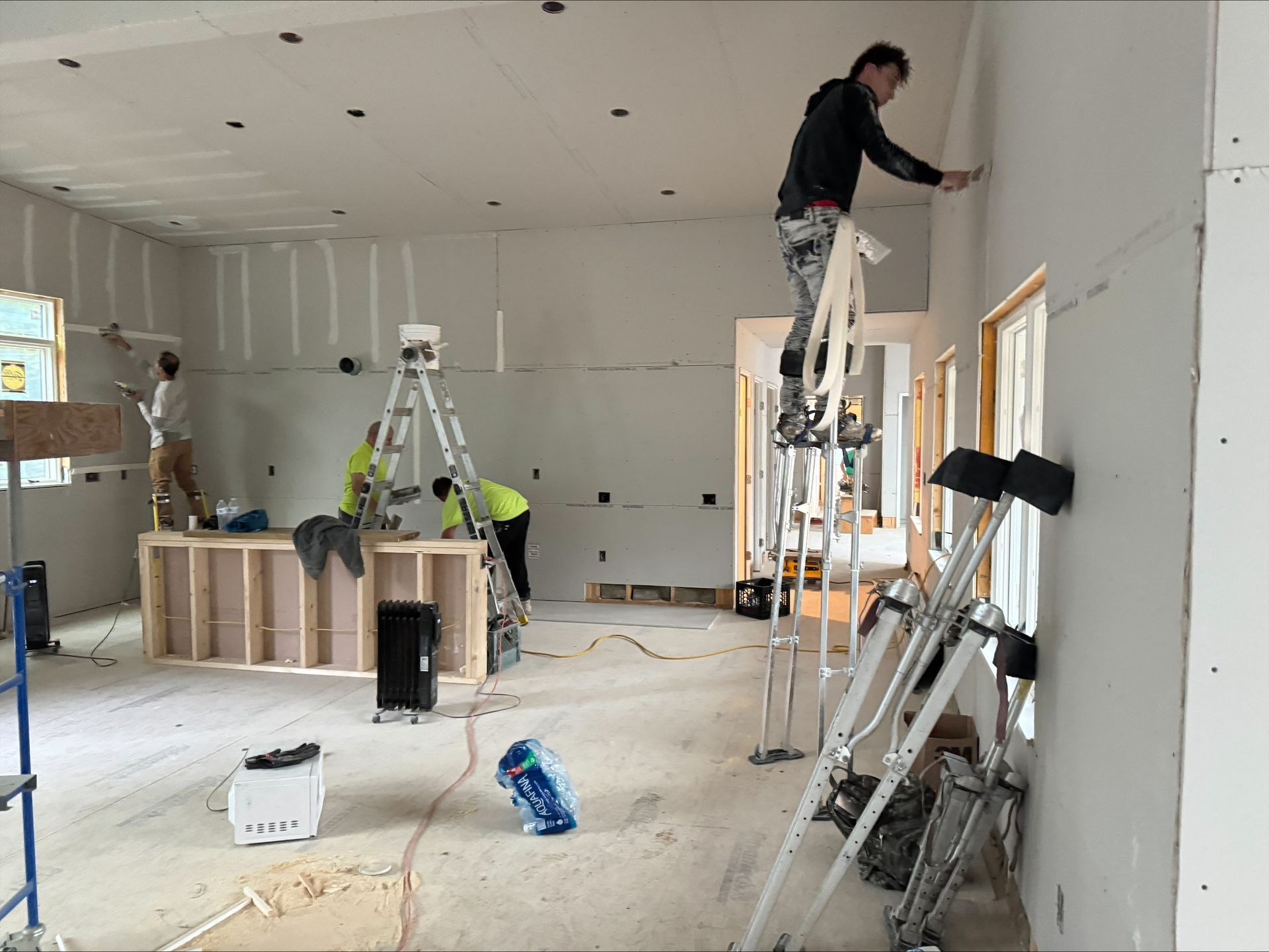 Two workers installing drywall in an unfinished room, one on a ladder near windows and tools on the floor