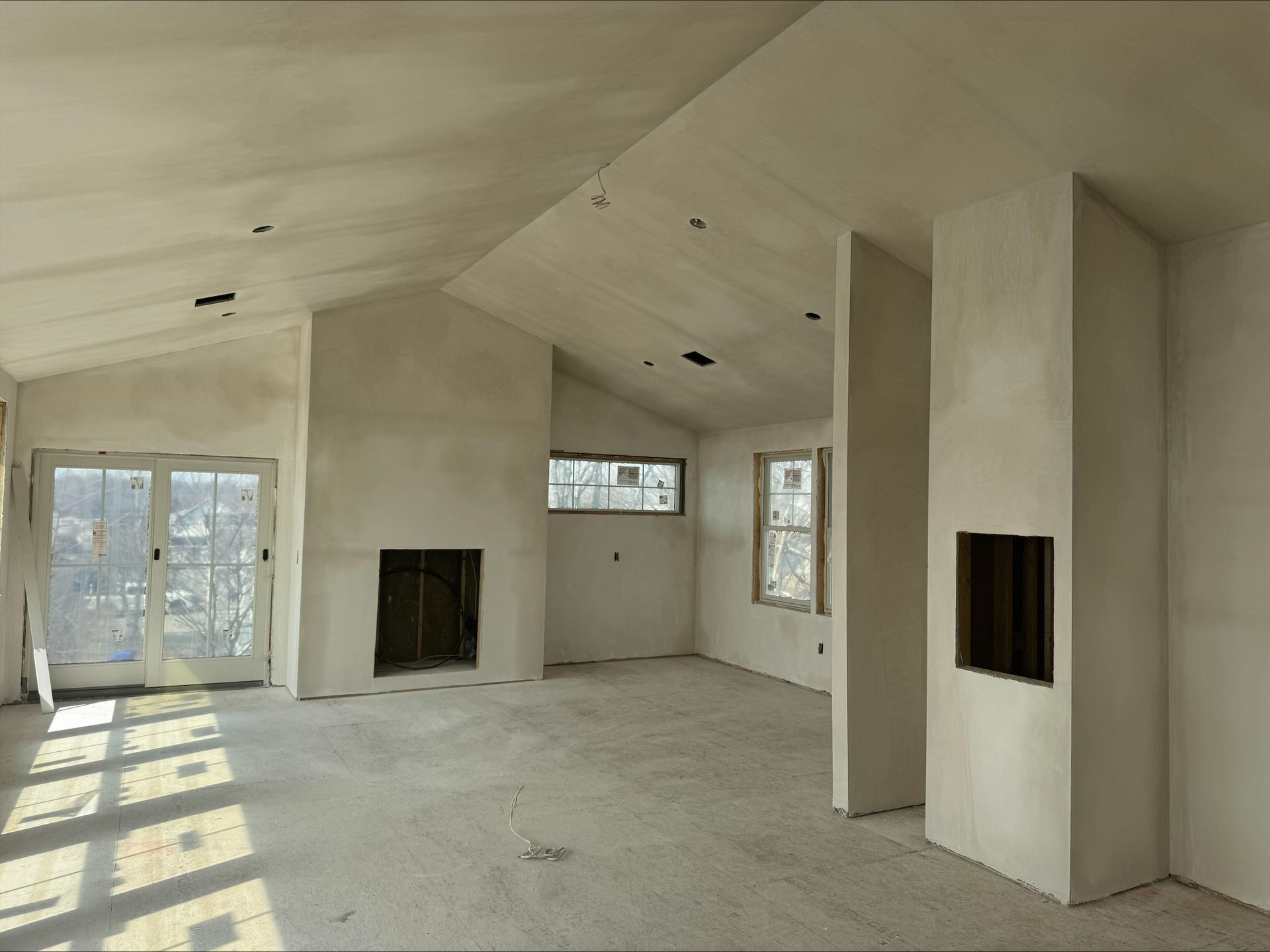 Empty unfinished room with white walls, fireplace openings, and sunlight streaming through windows and glass doors.