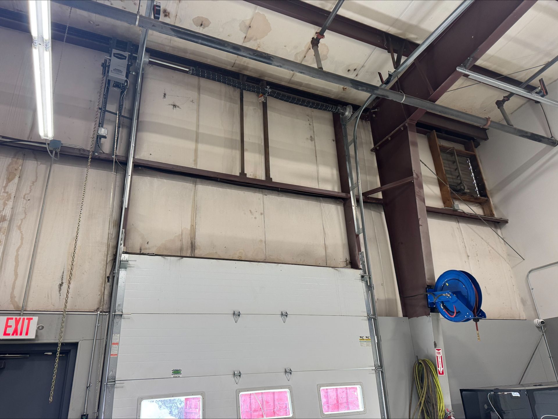 Interior of an industrial warehouse with a large roll-up door, exposed beams, and a blue wall-mounted fan.