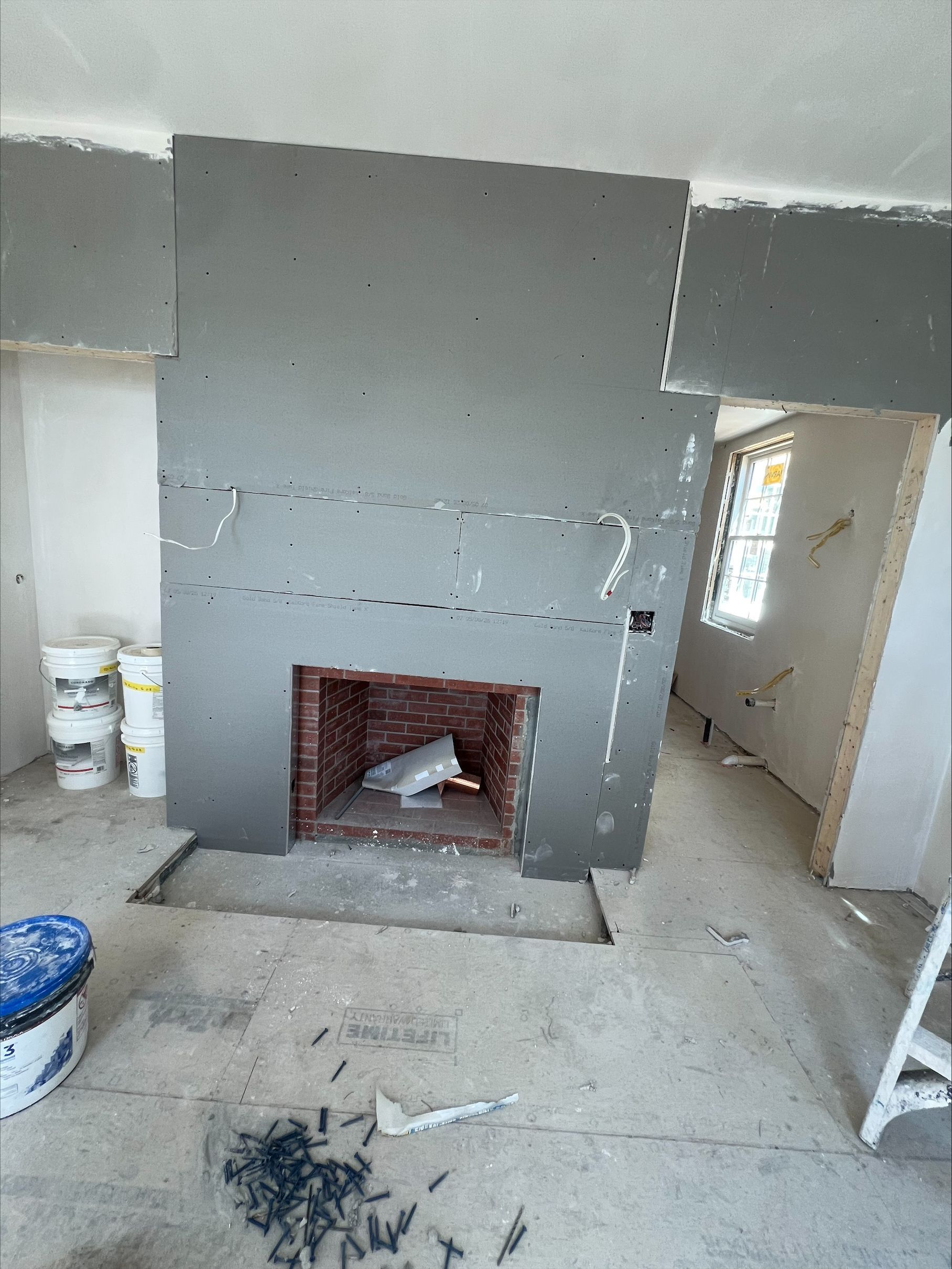 Partially finished room with gray drywall and a brick fireplace opening, construction tools and debris on the floor.