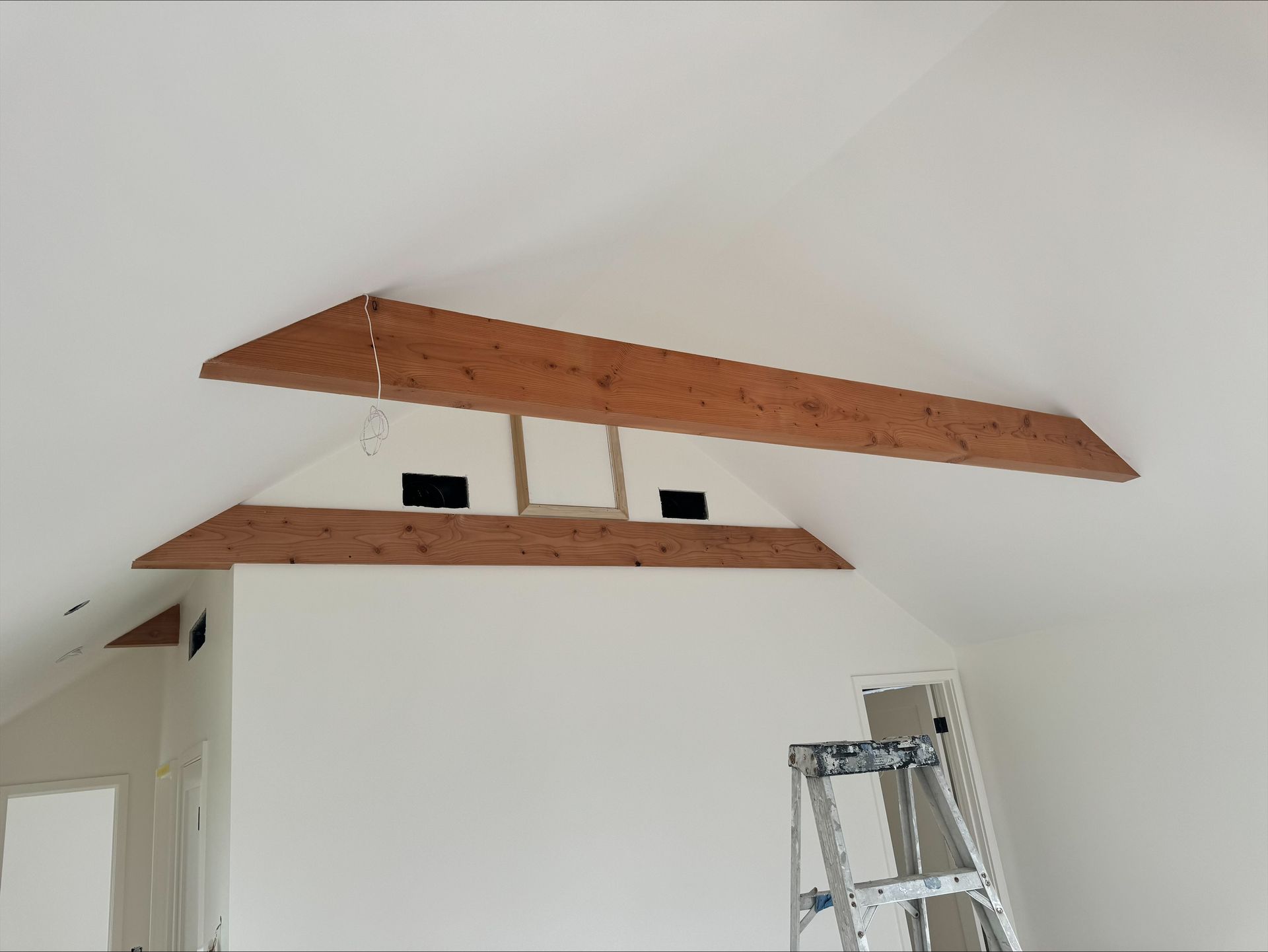 White room with two exposed wooden ceiling beams and a ladder below.