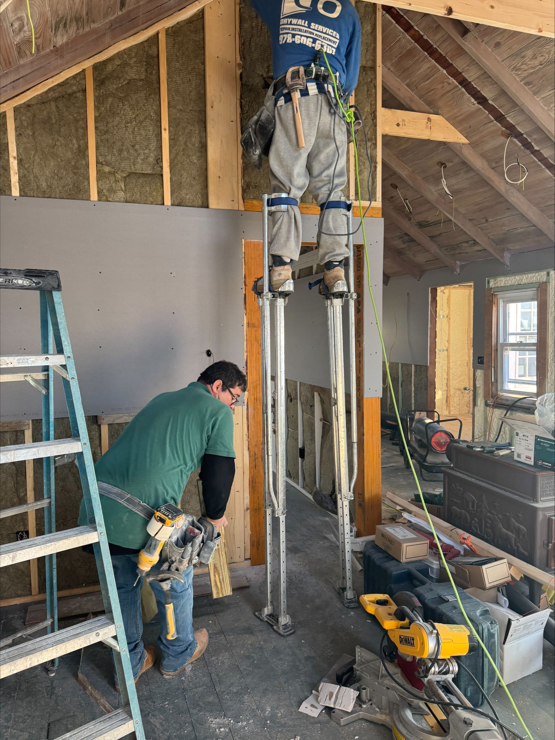Two workers installing ceiling insulation inside a partially finished room with ladders and tools.