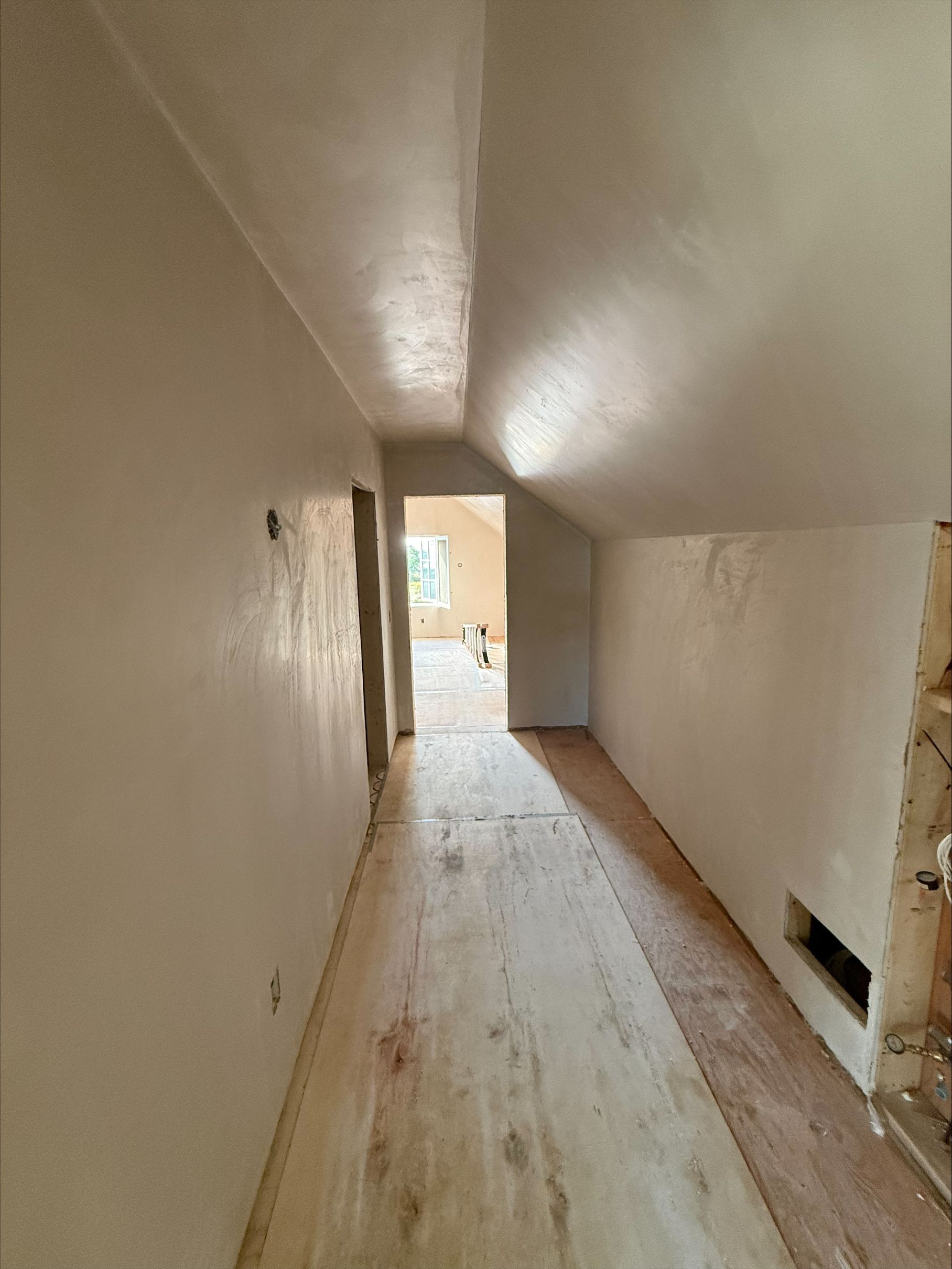 Narrow attic hallway with slanted ceiling, bare walls, and unfinished wooden floor leading to a bright doorway