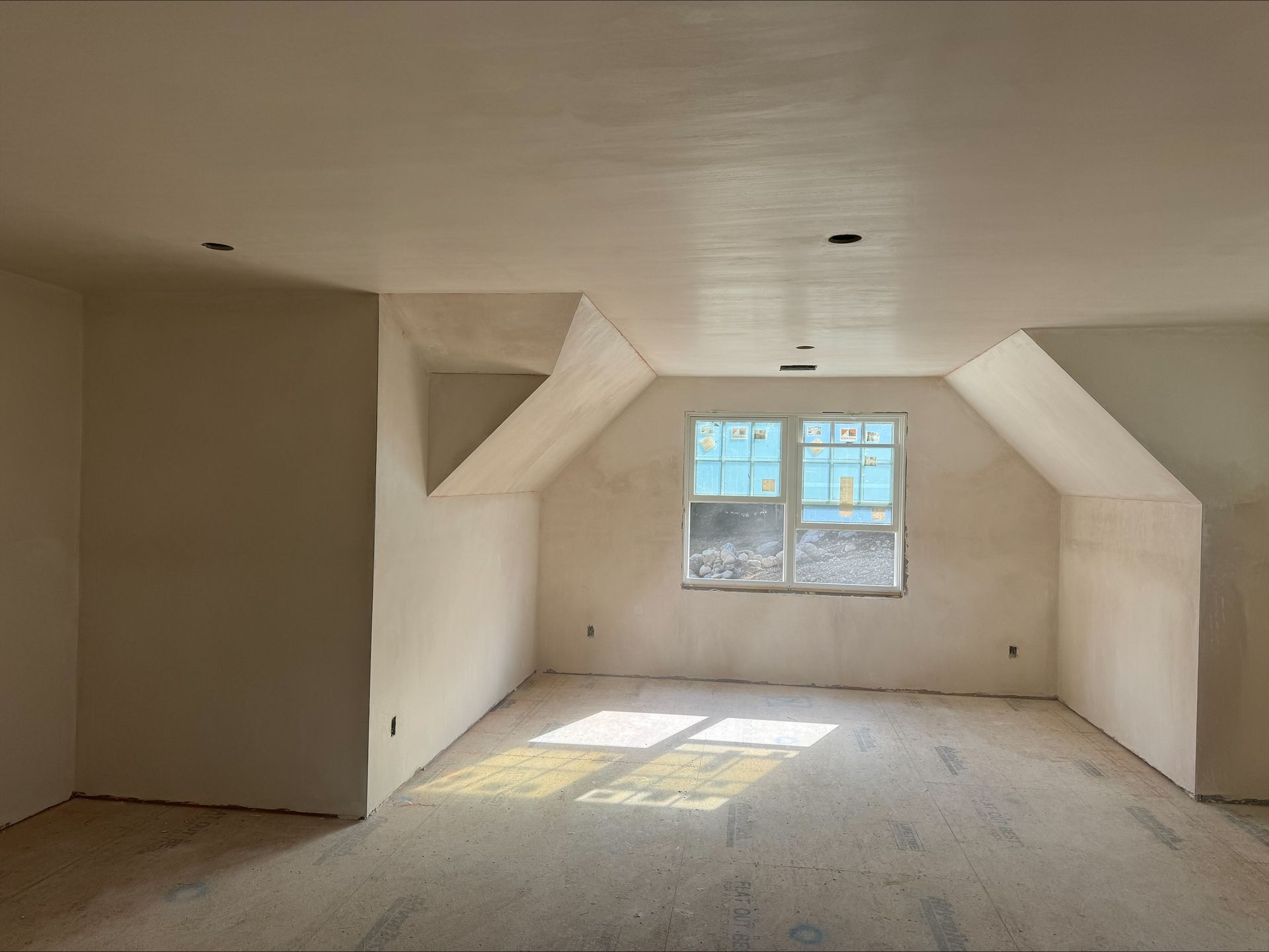 Empty unfinished attic room with sloped walls, bare floor, and sunlight through a small window