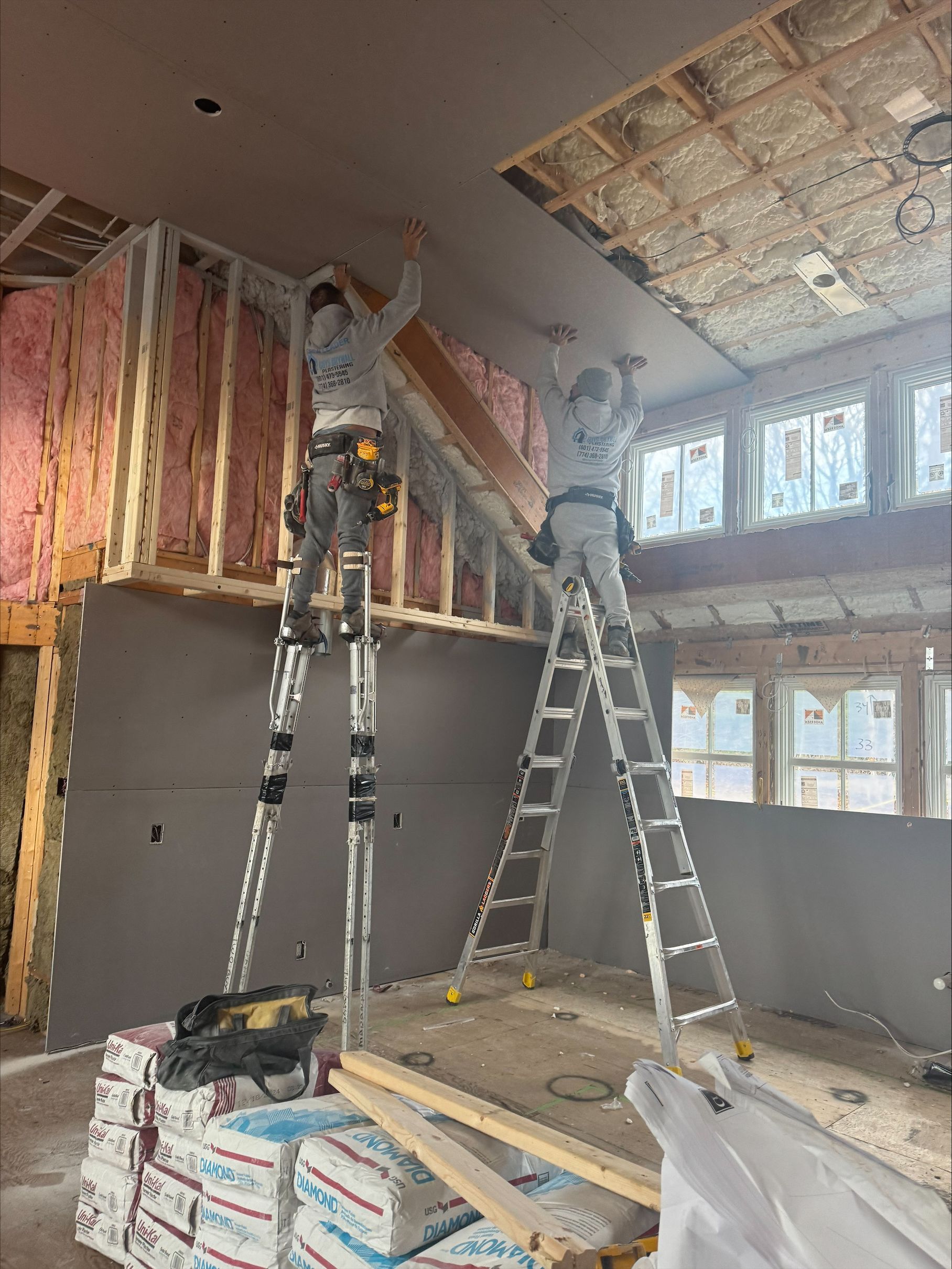 Workers installing insulation in a room with ladders, exposed walls, and construction materials