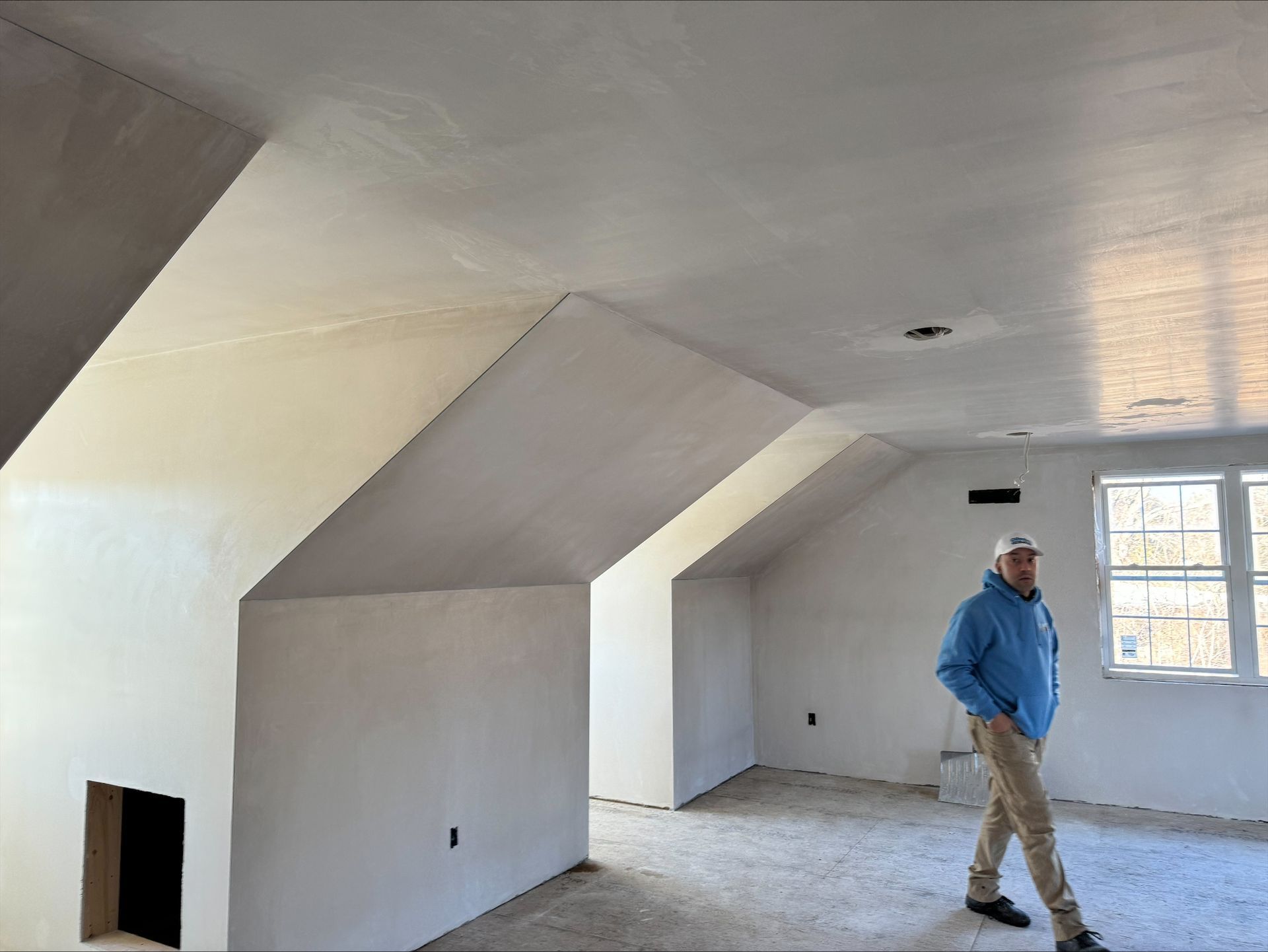 Unfinished attic room with white walls, sloped ceiling, and a person in a blue jacket standing near a window.