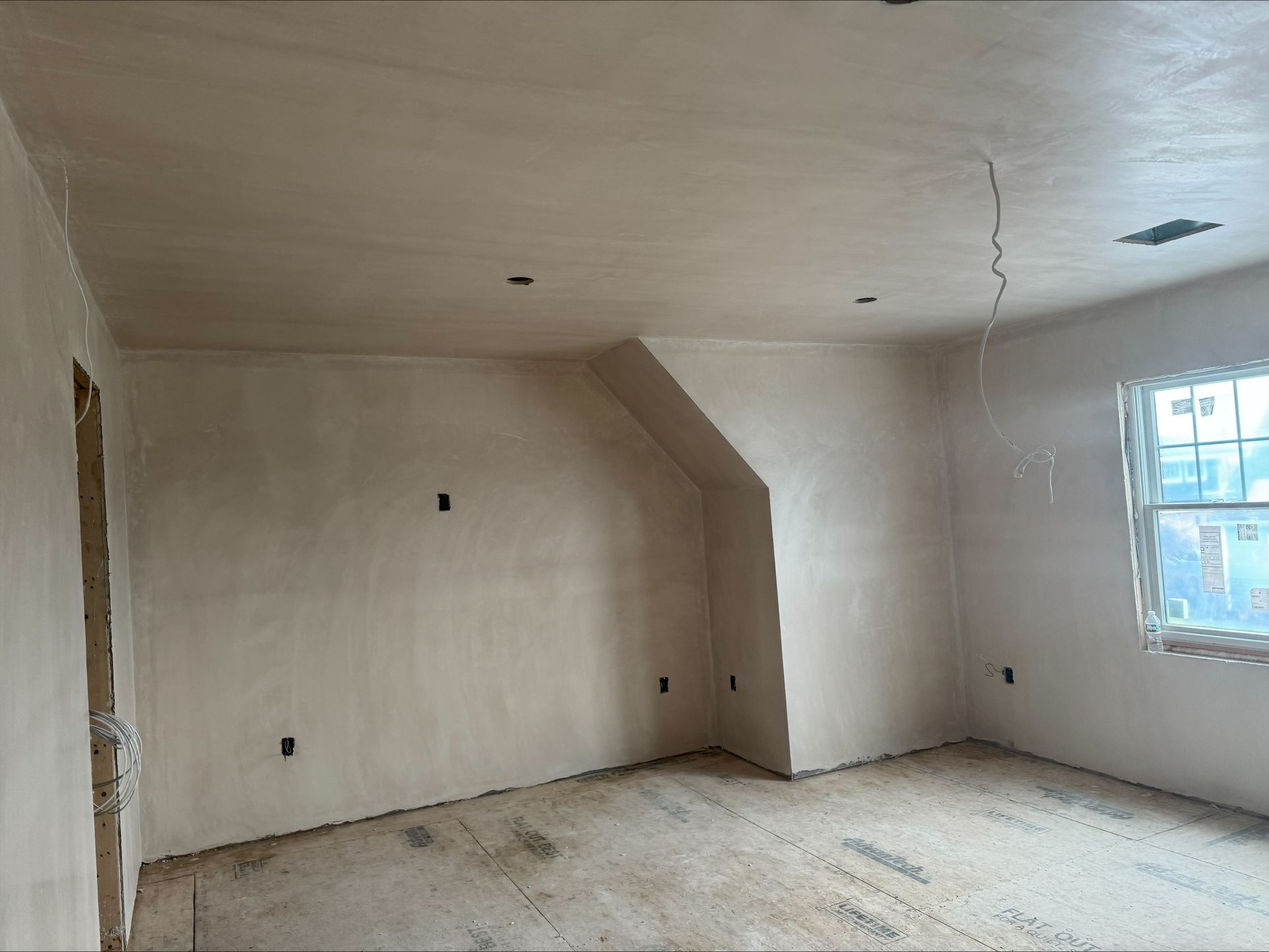 Empty unfinished room with white walls, bare floor, and a window letting in daylight