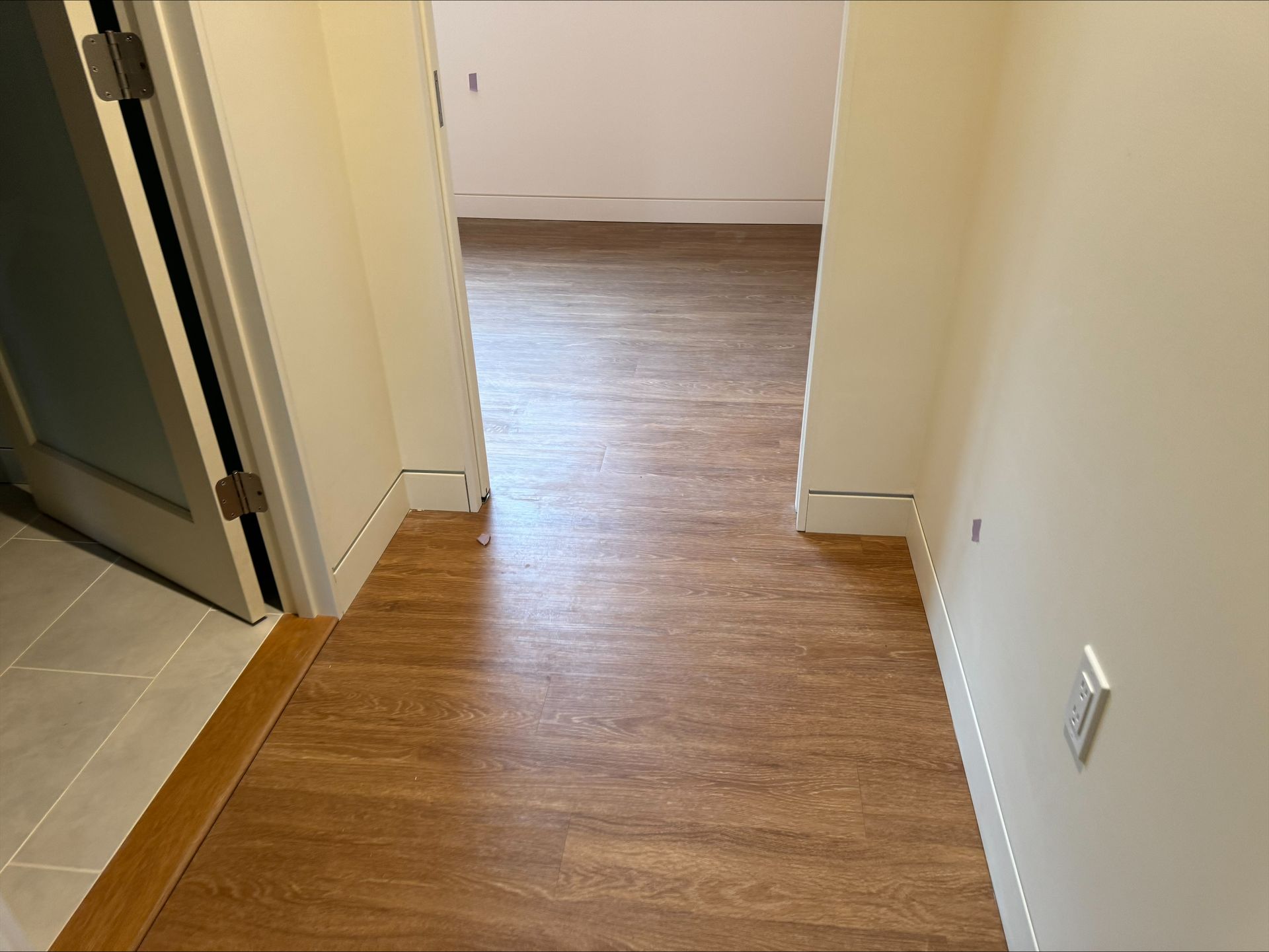 Empty hallway with wood-look flooring and white walls leading to a room.
