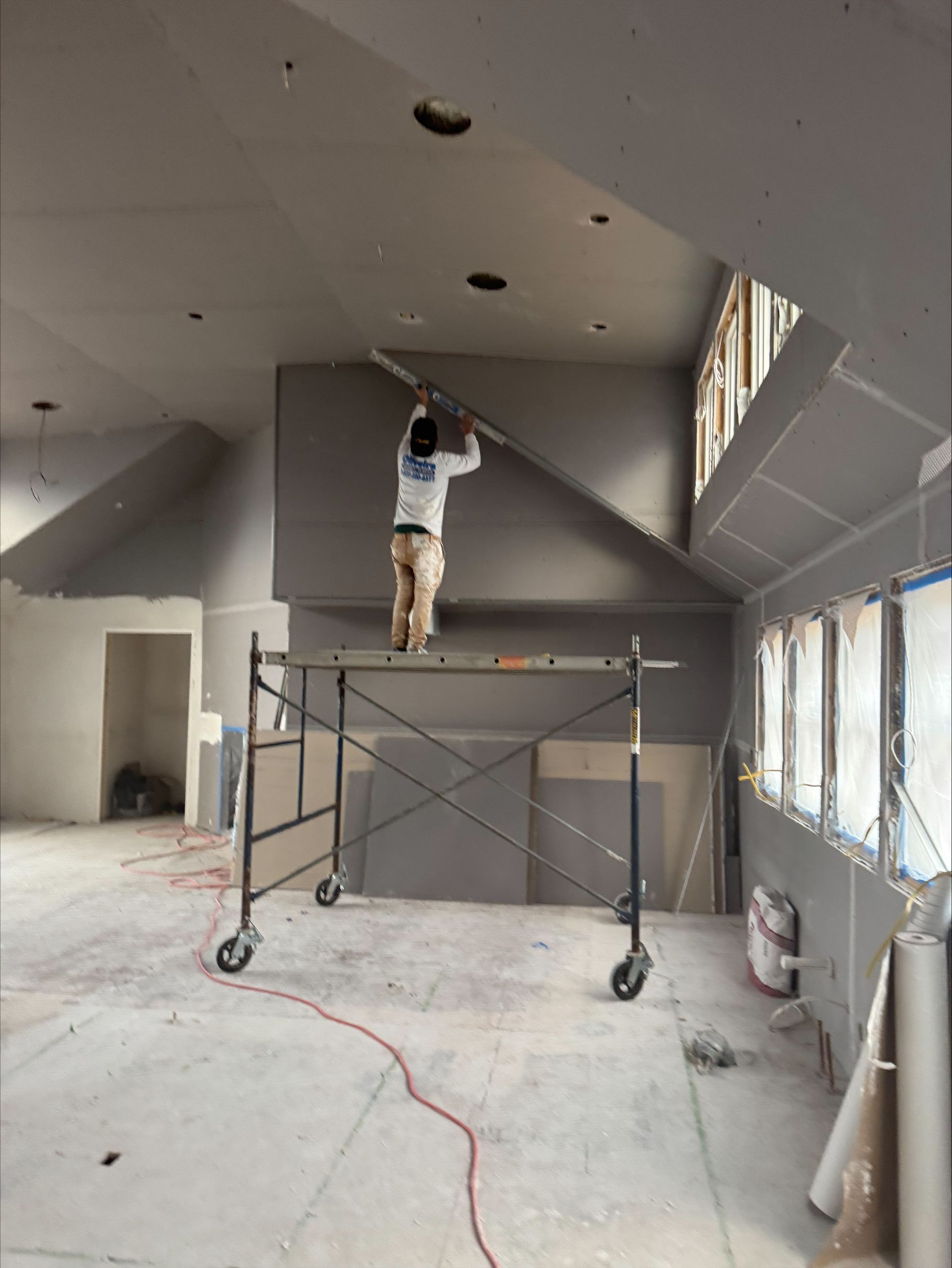 Worker on a scaffold painting a gray ceiling in an unfinished room with windows and construction tools.